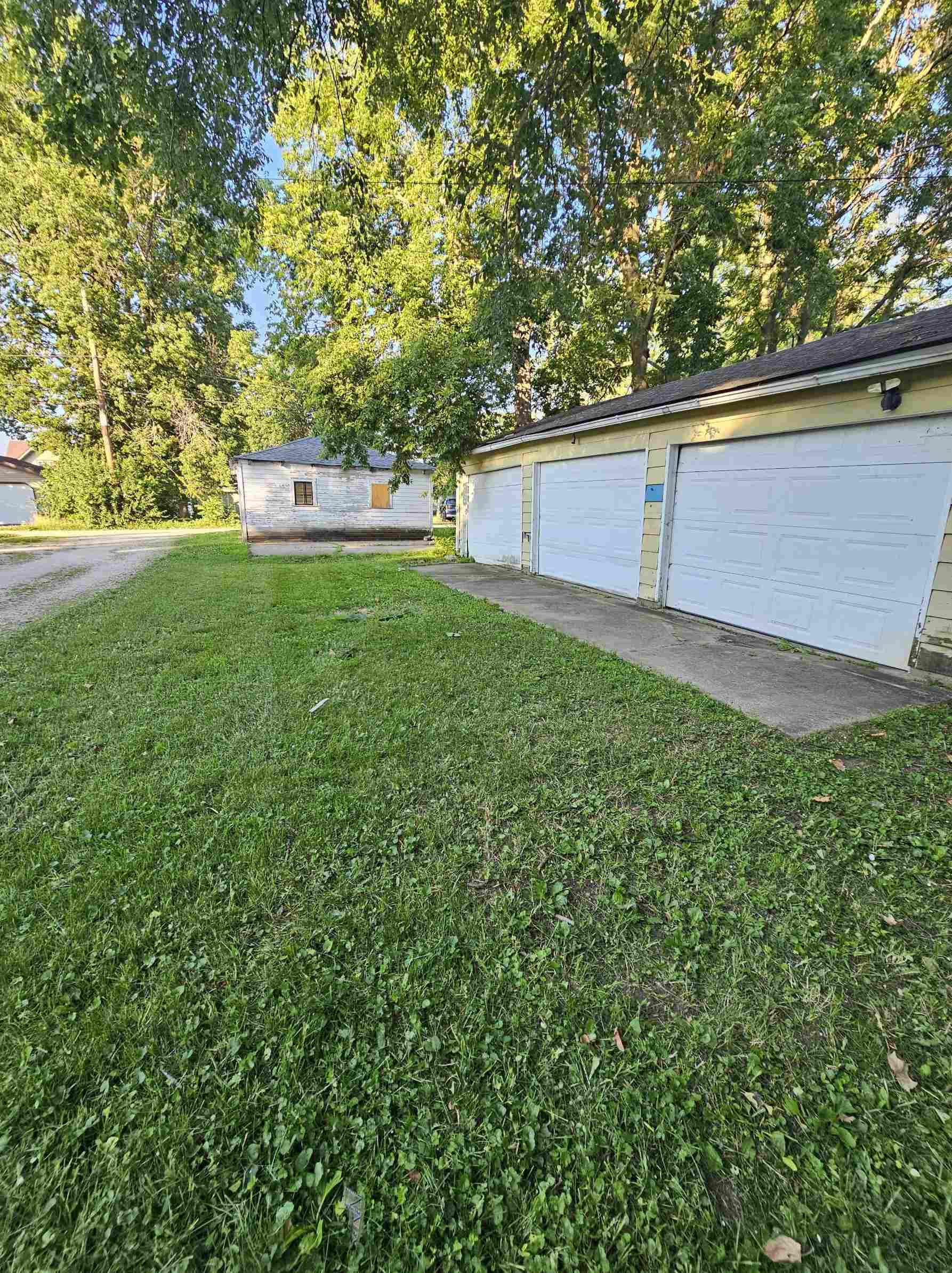 A grassy yard with a concrete driveway leading to a white garage with three doors, yellow siding, and a shingled roof. An older wooden building and tall green trees are in the background under a clear blue sky.