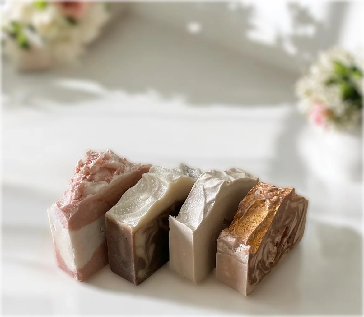 Four handmade soap bars with varying colors and patterns placed on a white surface, with blurred flowers in the background.