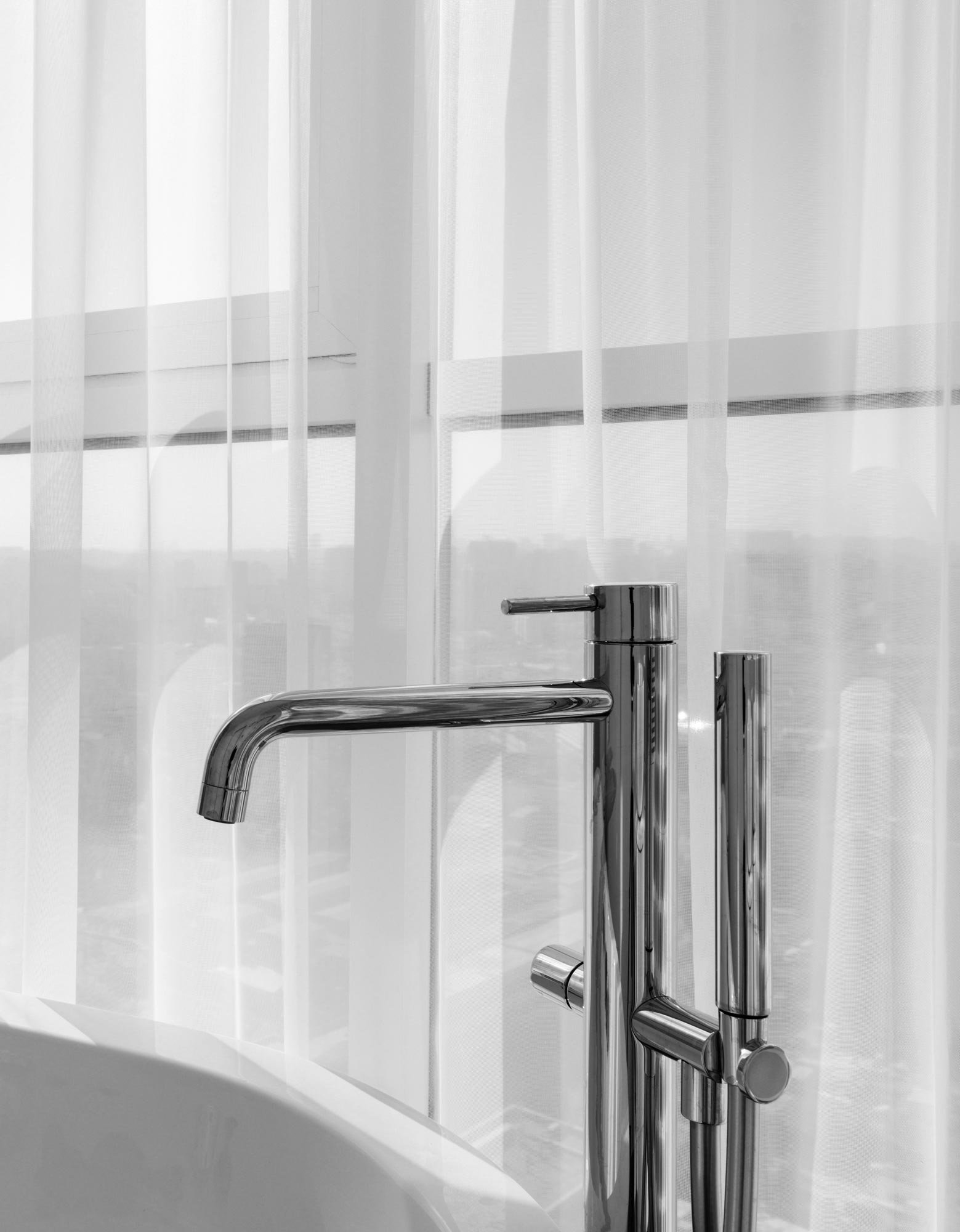 Close-up of a modern chrome bathroom faucet and handheld showerhead near a window with sheer curtains.