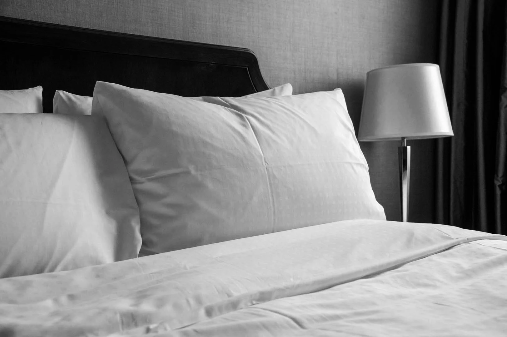 A neatly made bed with white pillows and bedding, next to a bedside lamp, in a hotel room, black and white photo.