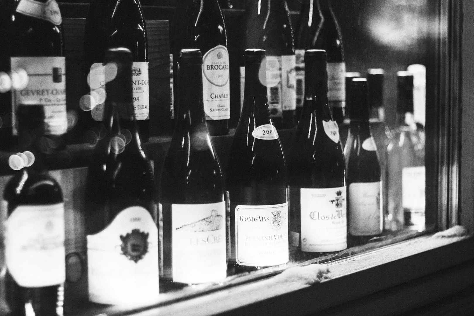A collection of wine bottles stored in a wine cooler or cellar with glass doors, with labels visible on some bottles, and some bottles having tags with numbers.
