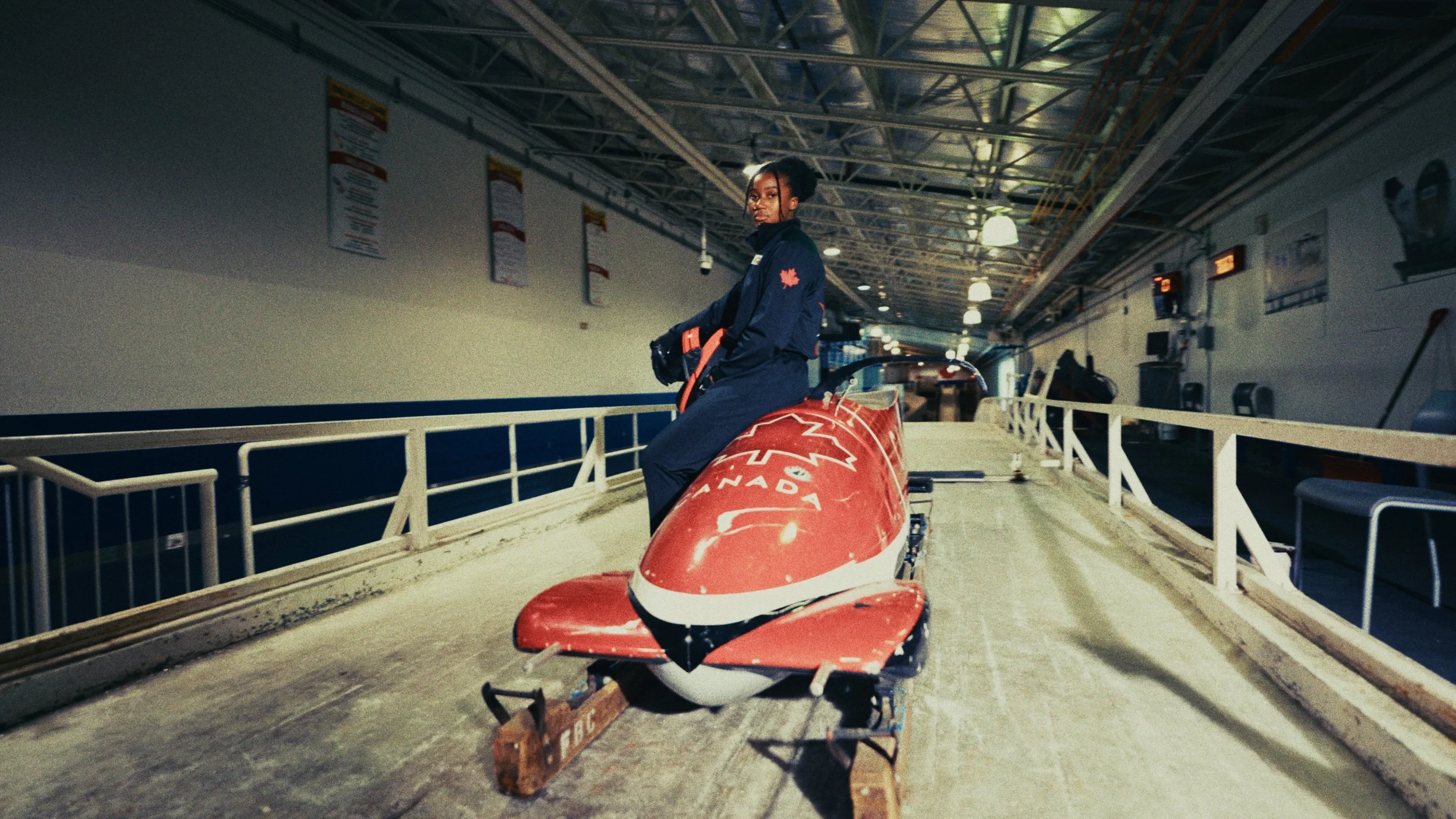 An athlete in a navy jacket and pants sitting on a red bobsled on an indoor ice rink track.