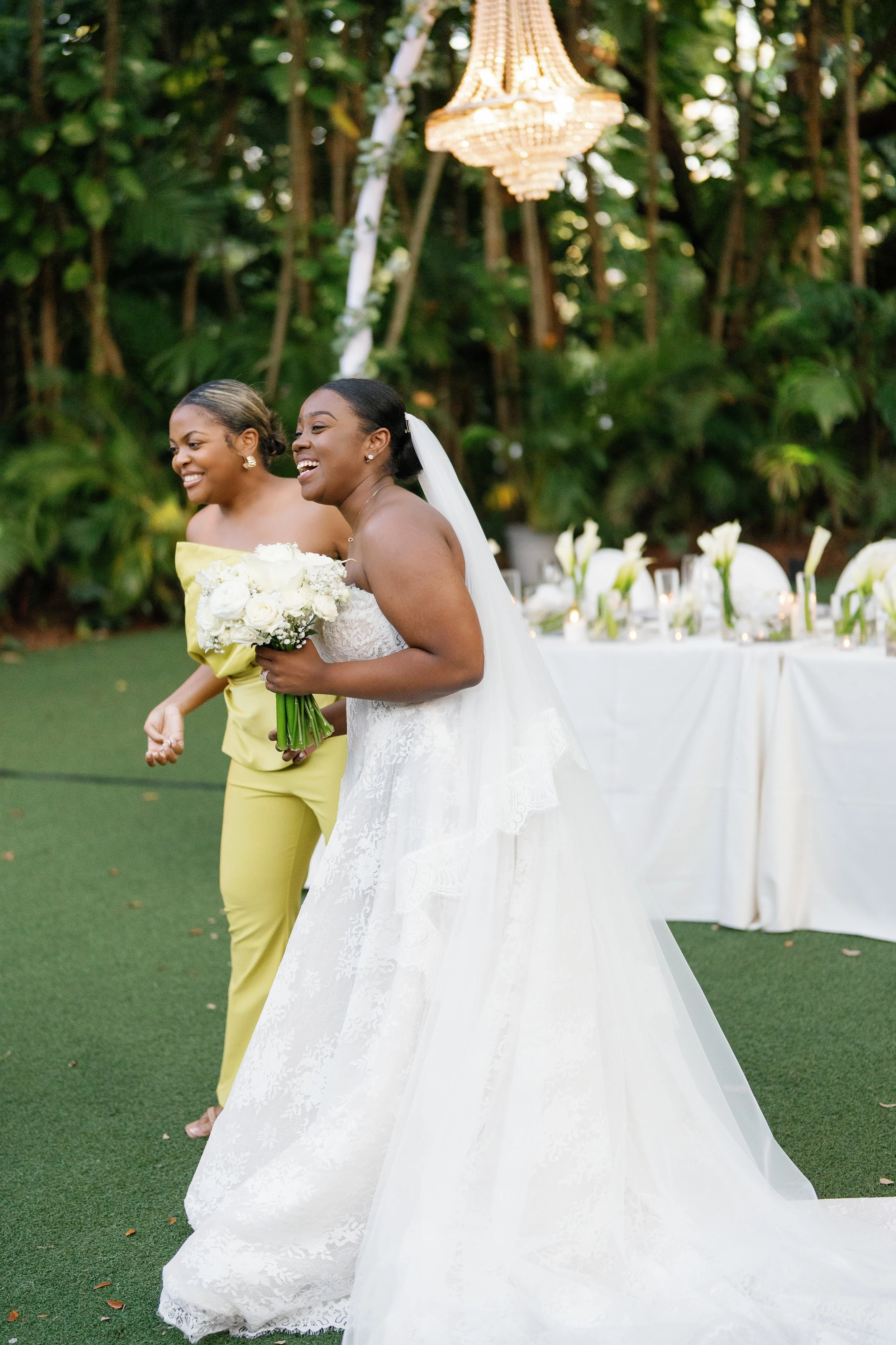 Two women, one in a white wedding gown with a veil and the other in a yellow outfit, laughing and holding a bouquet of white flowers at an outdoor wedding reception with a decorated table and a chandelier overhead.