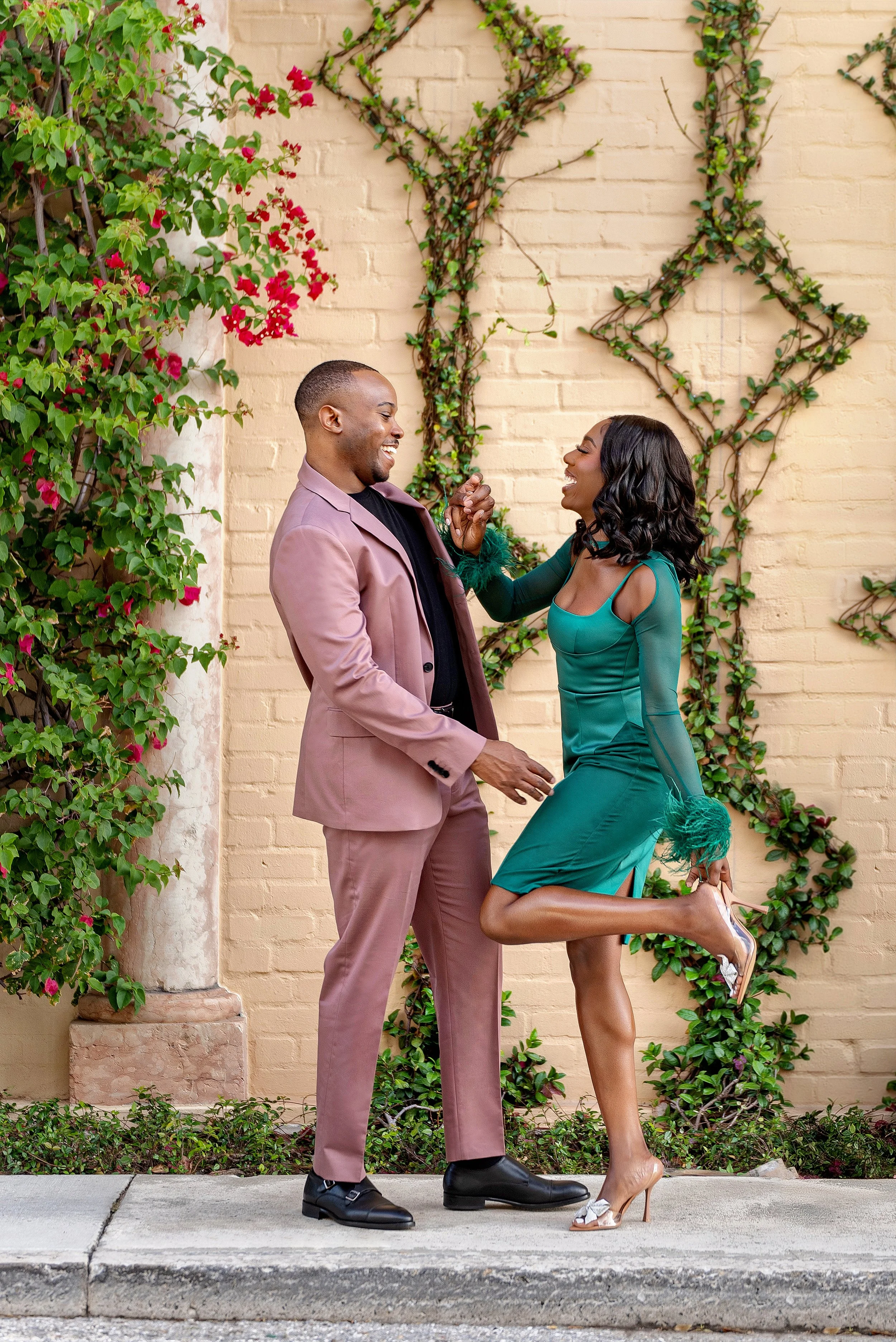 A joyful couple dancing and laughing on a sidewalk in front of a decorated brick wall with green vines and flowers.