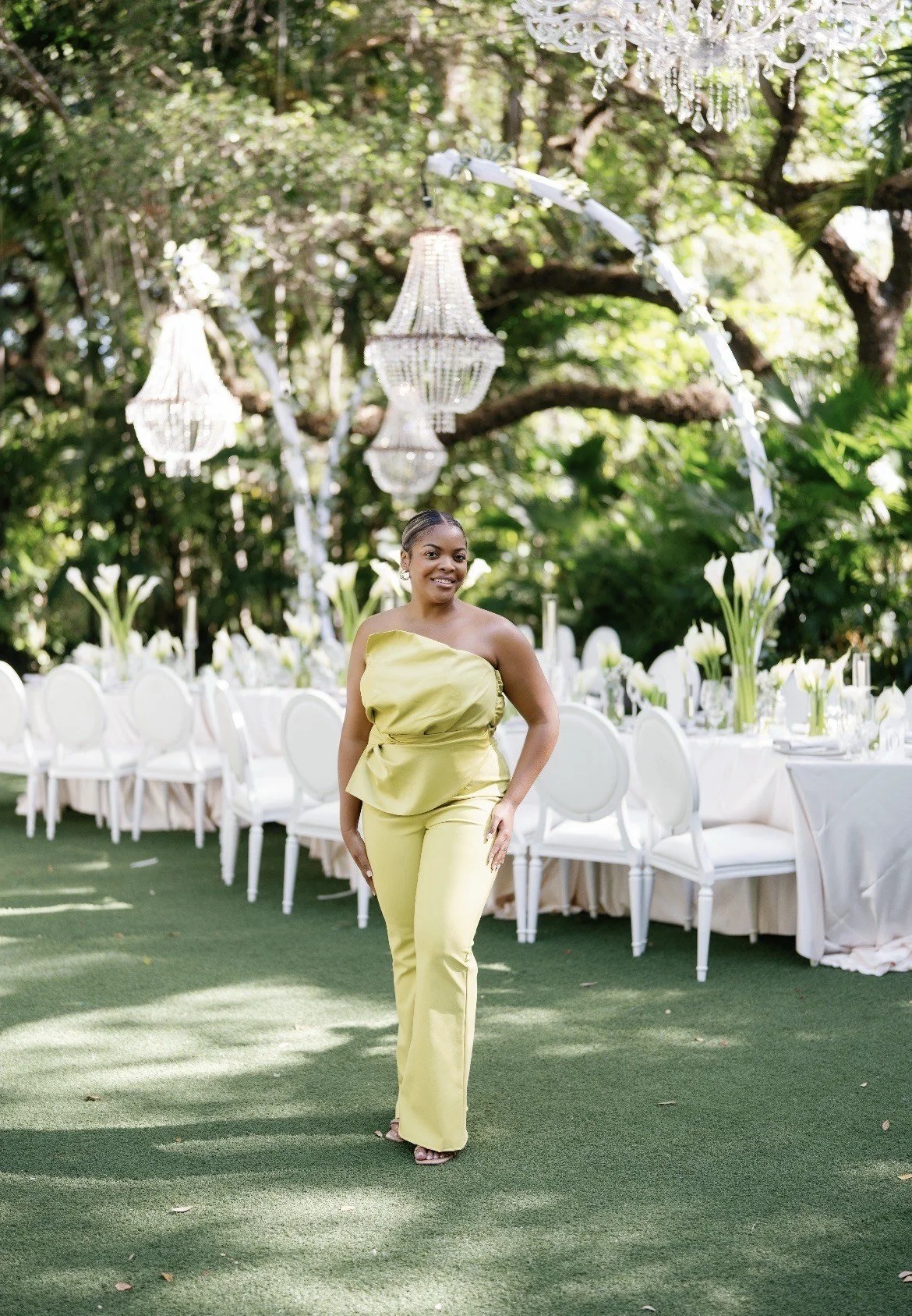 A smiling woman in a yellow off-the-shoulder jumpsuit standing in front of a decorated outdoor event table with white chairs, tall white floral arrangements, and hanging chandeliers amidst greenery.