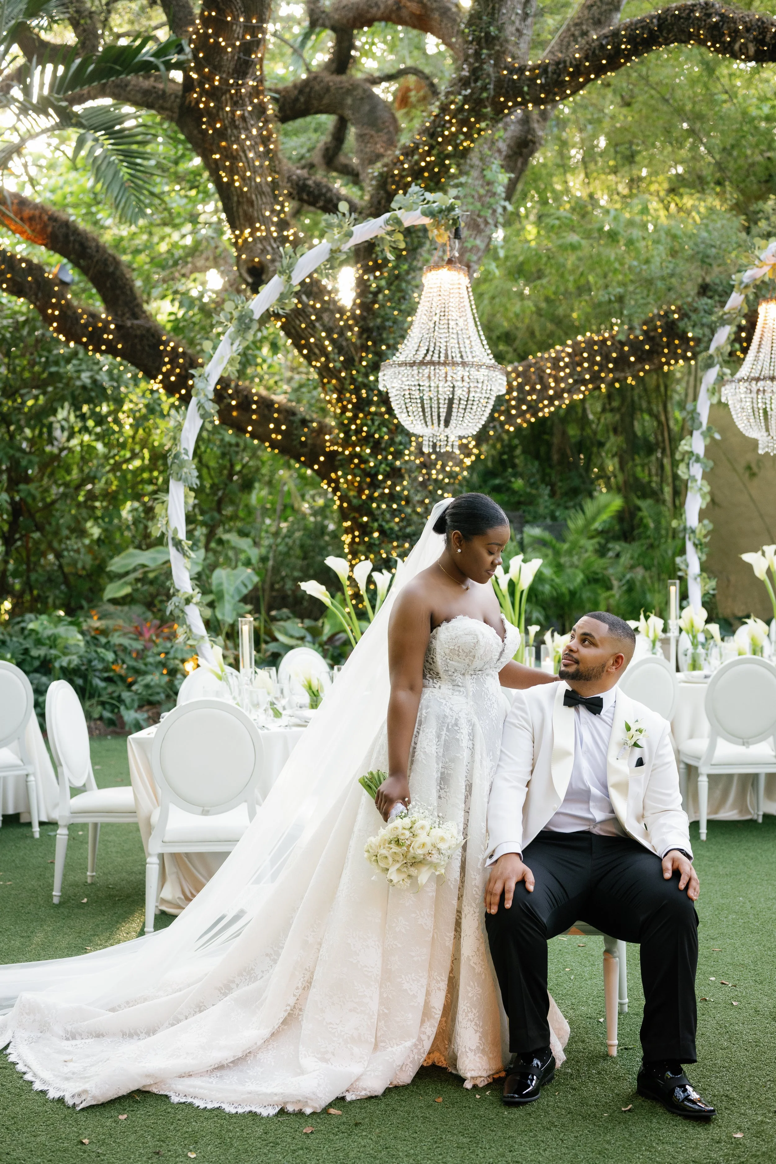 A bride and groom in wedding attire sharing a moment outdoors under a decorated arch with romantic lighting and chandeliers, surrounded by white chairs and a table setting in a lush garden.
