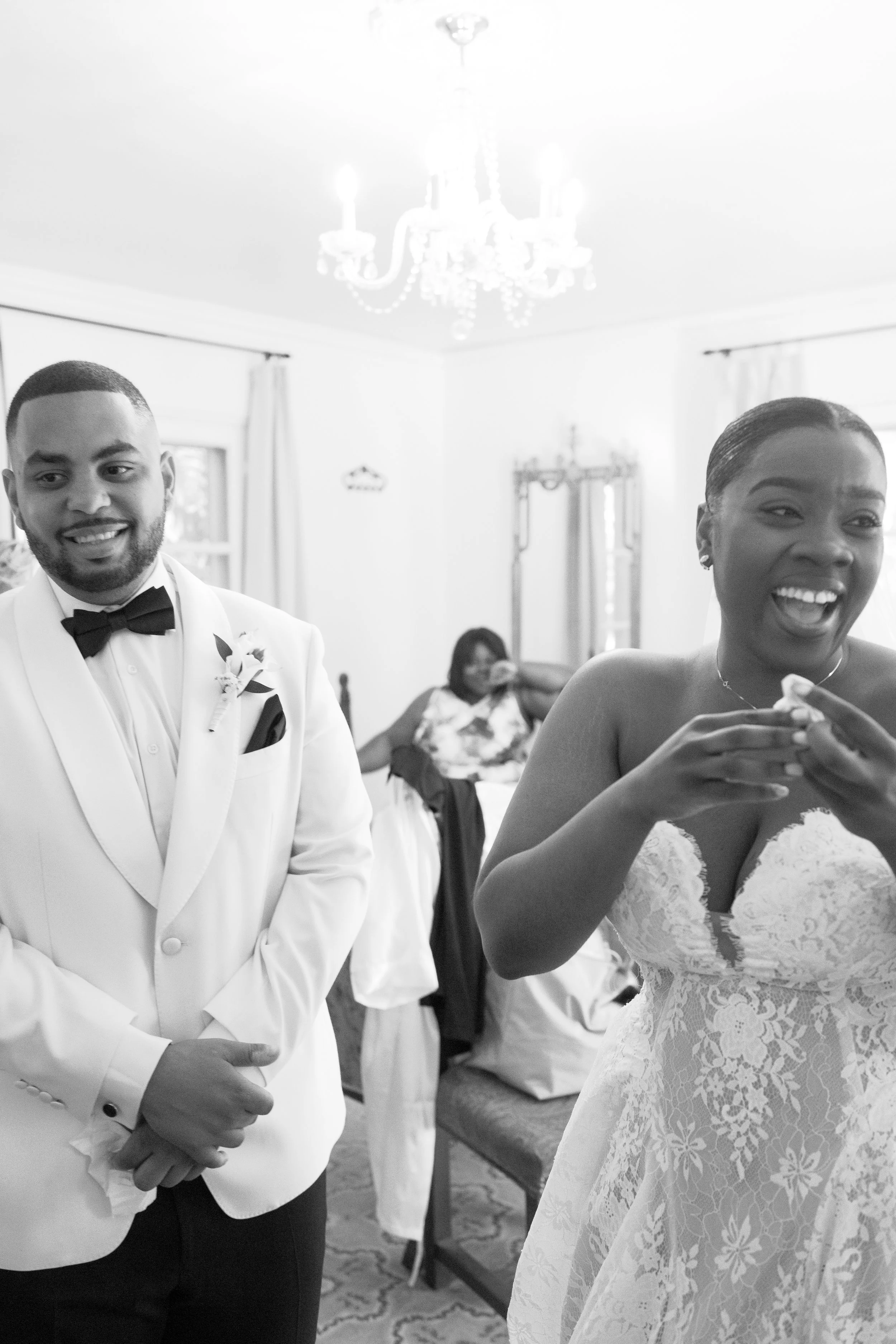 A bride and groom on their wedding day, smiling in a room with a chandelier and another woman in the background smiling.