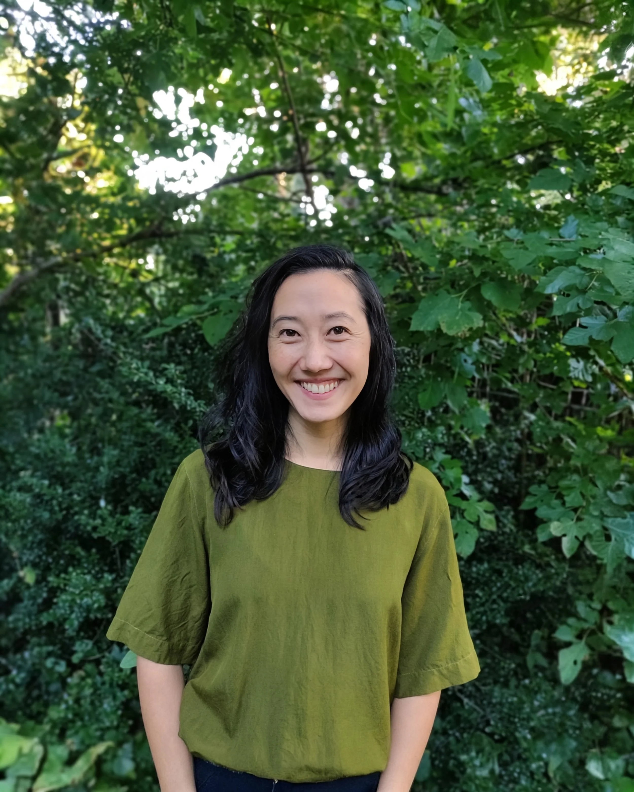 Image of Katie Chen: Woman with dark hair wearing an olive green top, smiling outdoors in front of lush green foliage.