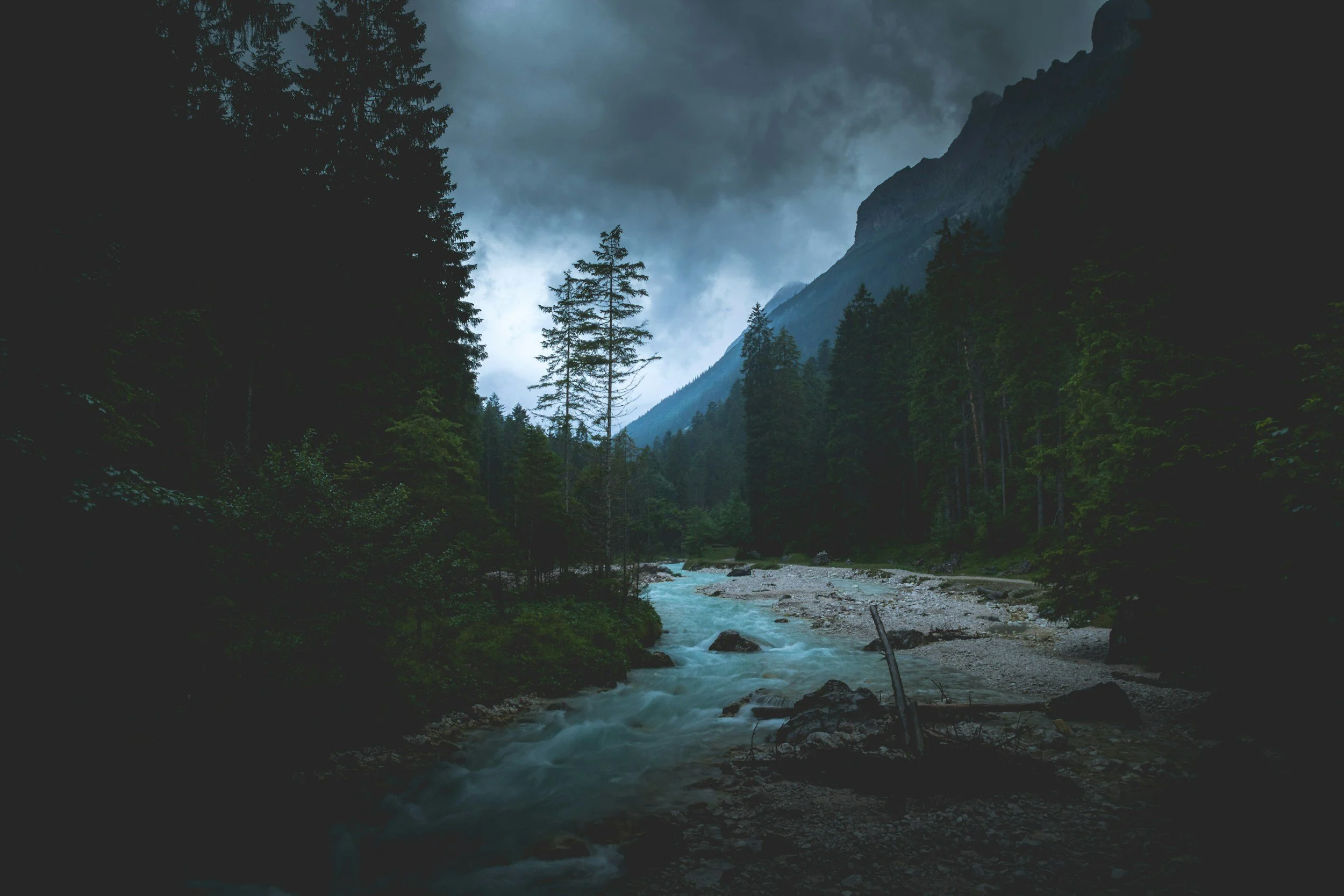 A mountain river flowing through a dense evergreen forest with cloudy skies overhead.