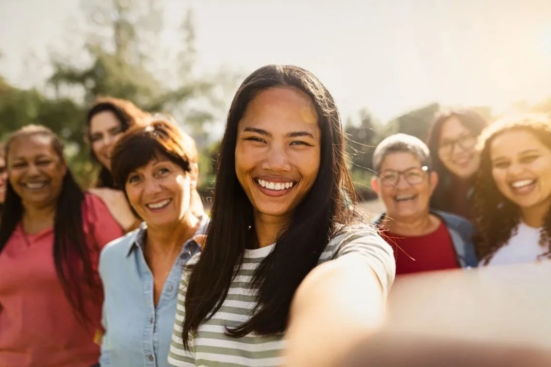 A group of diverse women smiling for a selfie outdoors during daytime.