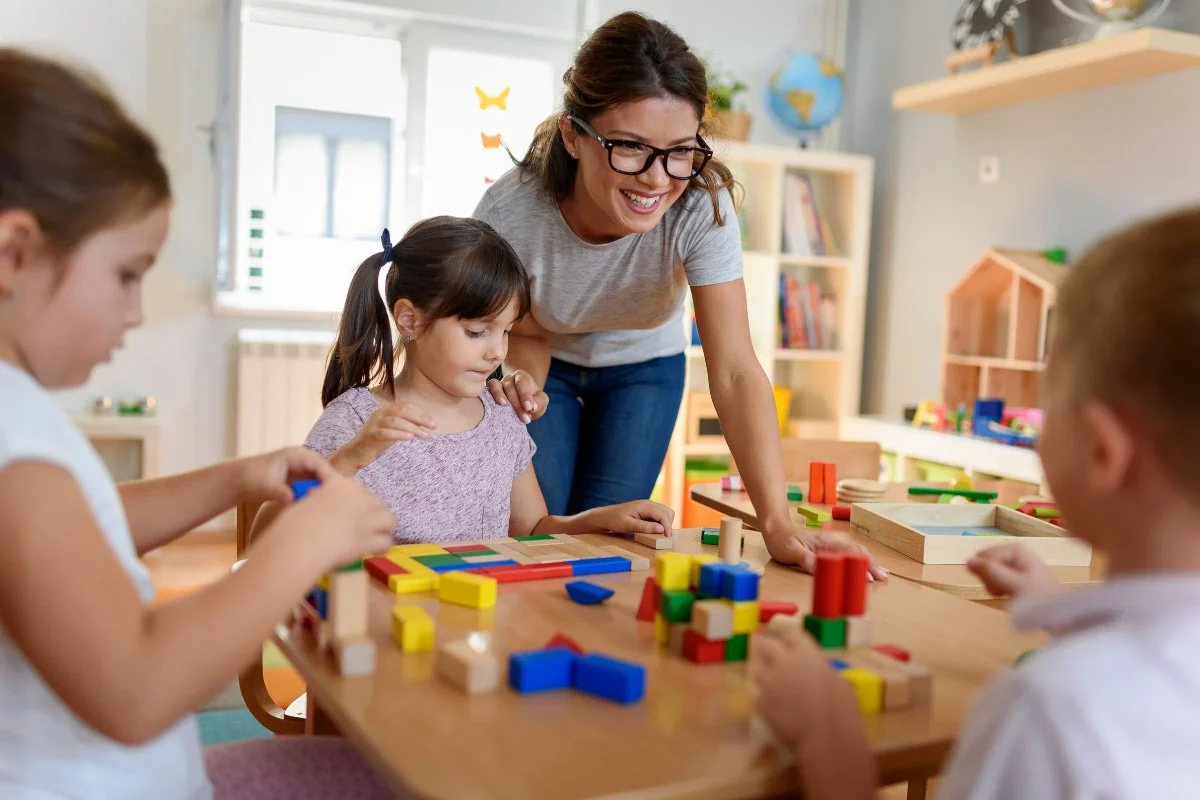 Teacher and young children playing with colorful wooden building blocks at a table in a classroom.