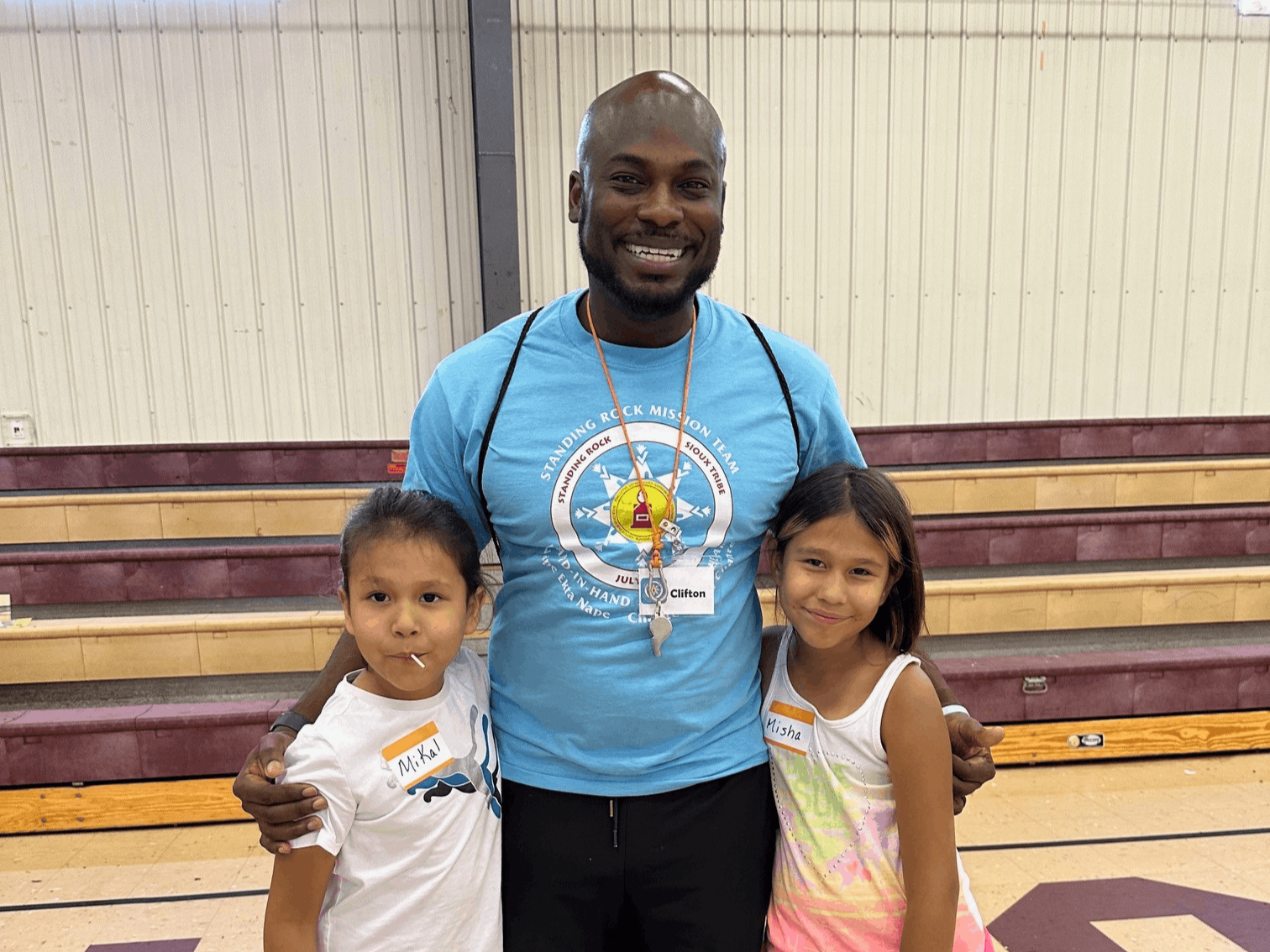 A man standing in a gymnasium with two young girls, one with a lollipop and the other with a smile, all smiling at the camera.