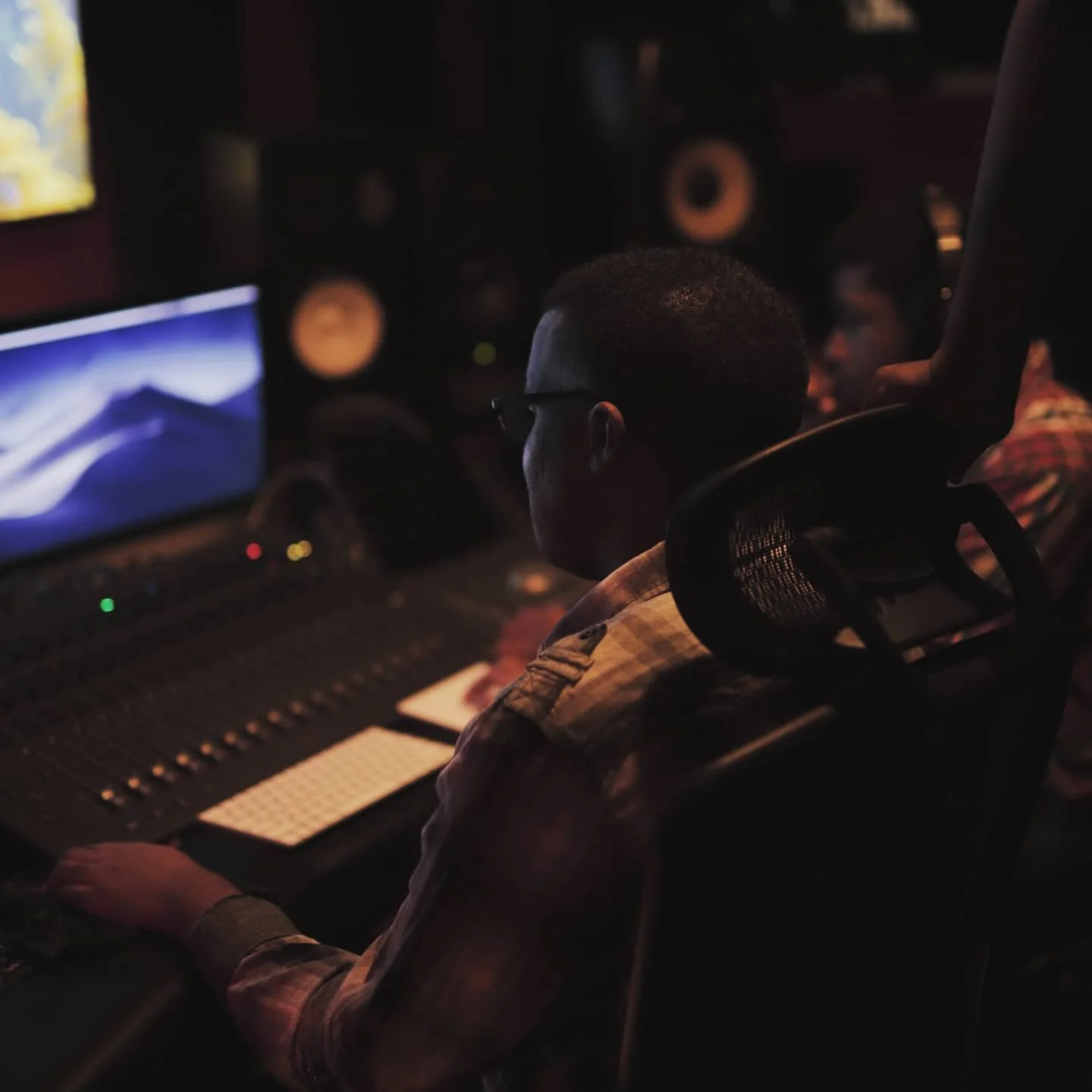 A person with glasses working on audio mixing in a dark recording studio with speakers and a computer monitor.
