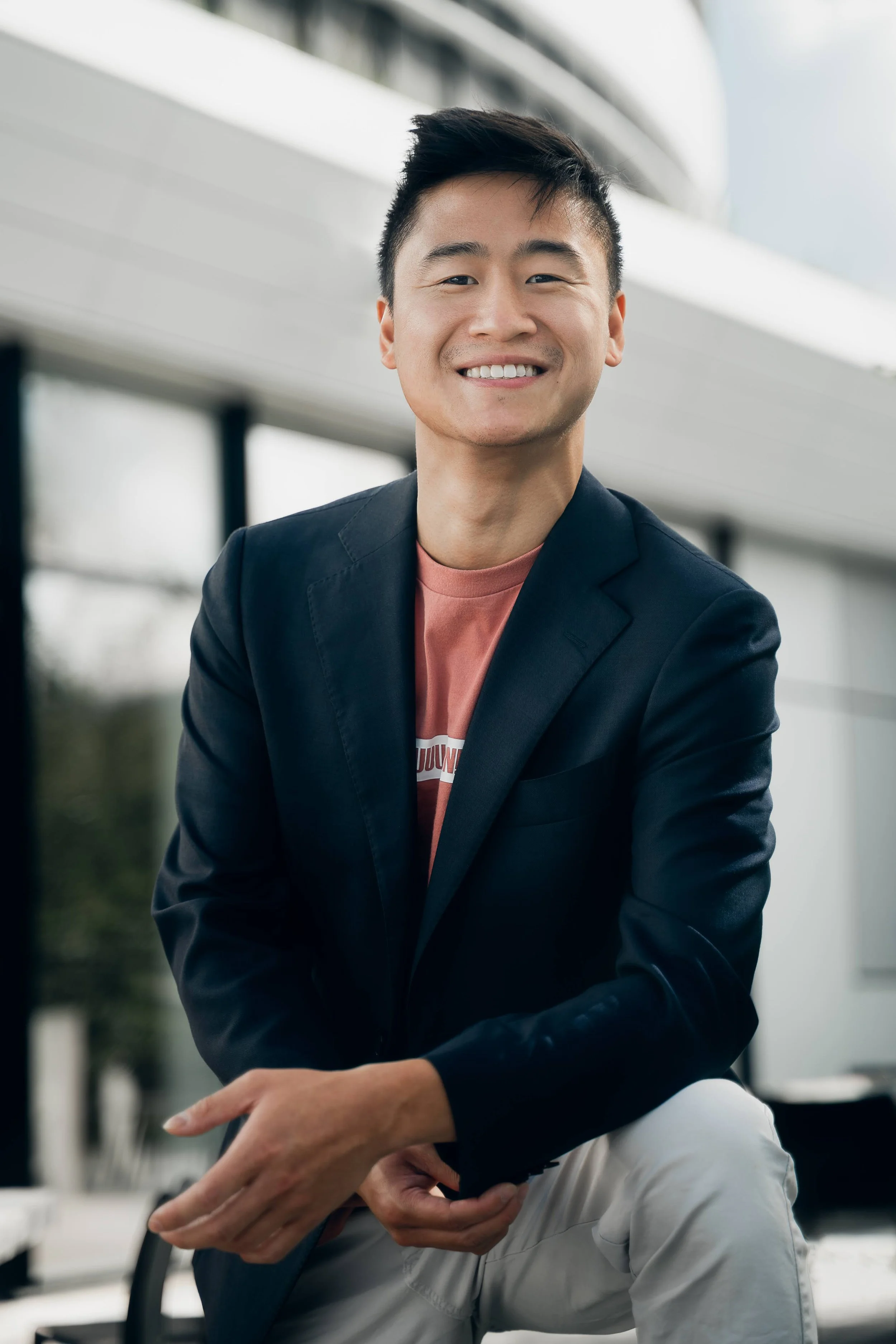 Young Asian man smiling outdoors, wearing a dark blazer over a pink t-shirt with text, and light-colored pants, leaning on a surface with one leg raised. Hao Wu Personal Blog.