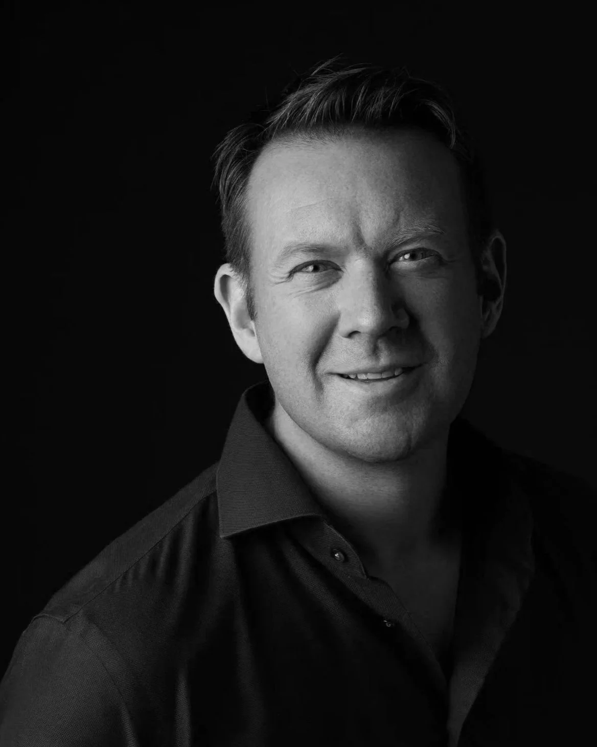 Black and white portrait of a smiling man with short, styled hair, wearing a collared shirt against a dark background.