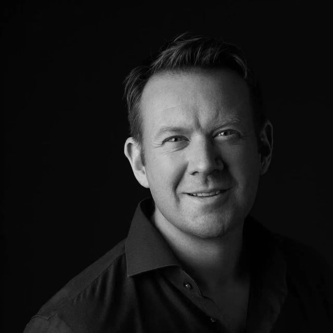 Black and white portrait of a smiling man with short, styled hair, wearing a dark collared shirt, against a dark background.