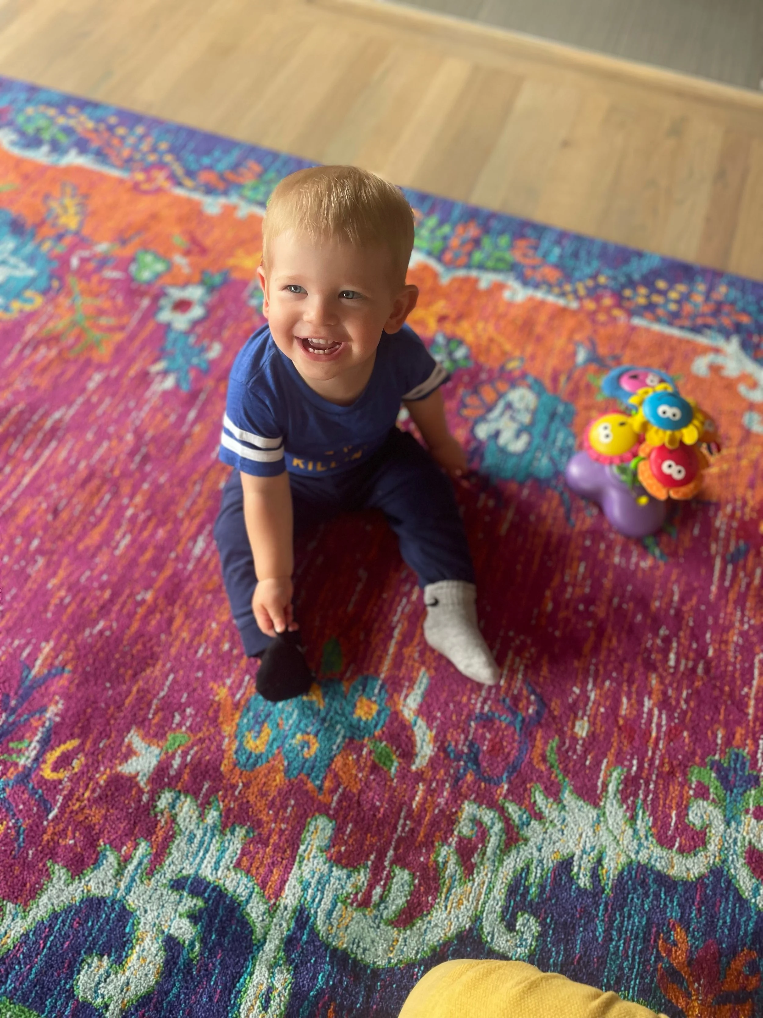 A young boy with short blond hair, wearing a blue shirt and dark pants, is sitting on a colorful, patterned rug, smiling and looking up. Beside him is a purple toy with several small, round, colorful plush toys attached.