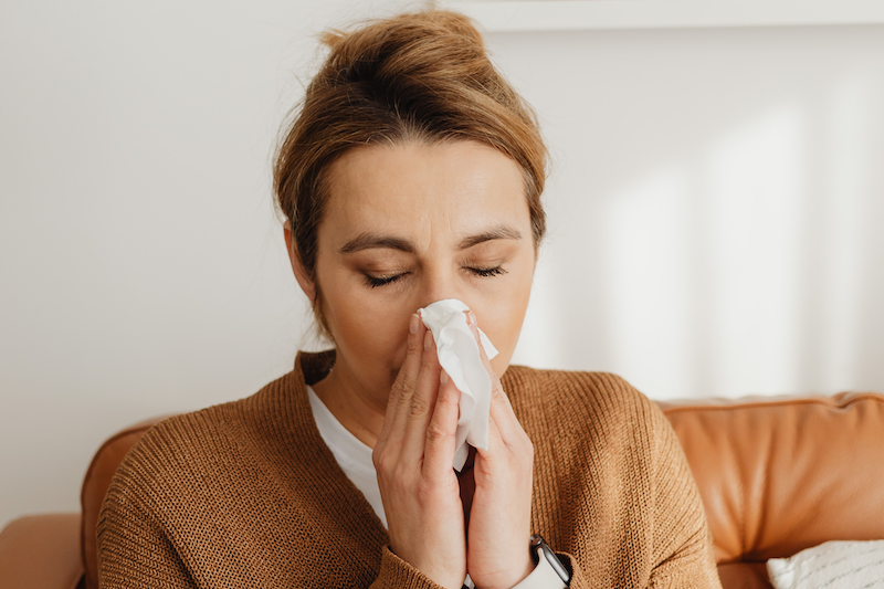 A woman sitting on a leather couch, sneezing or blowing her nose into a tissue.