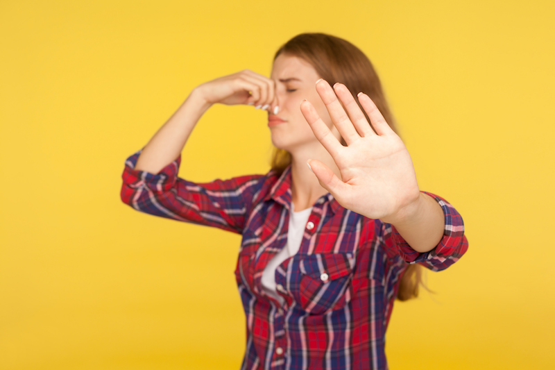 Woman with red hair wearing a red plaid shirt pressing her finger to her nose and holding up her hand in a stop gesture against a yellow background.