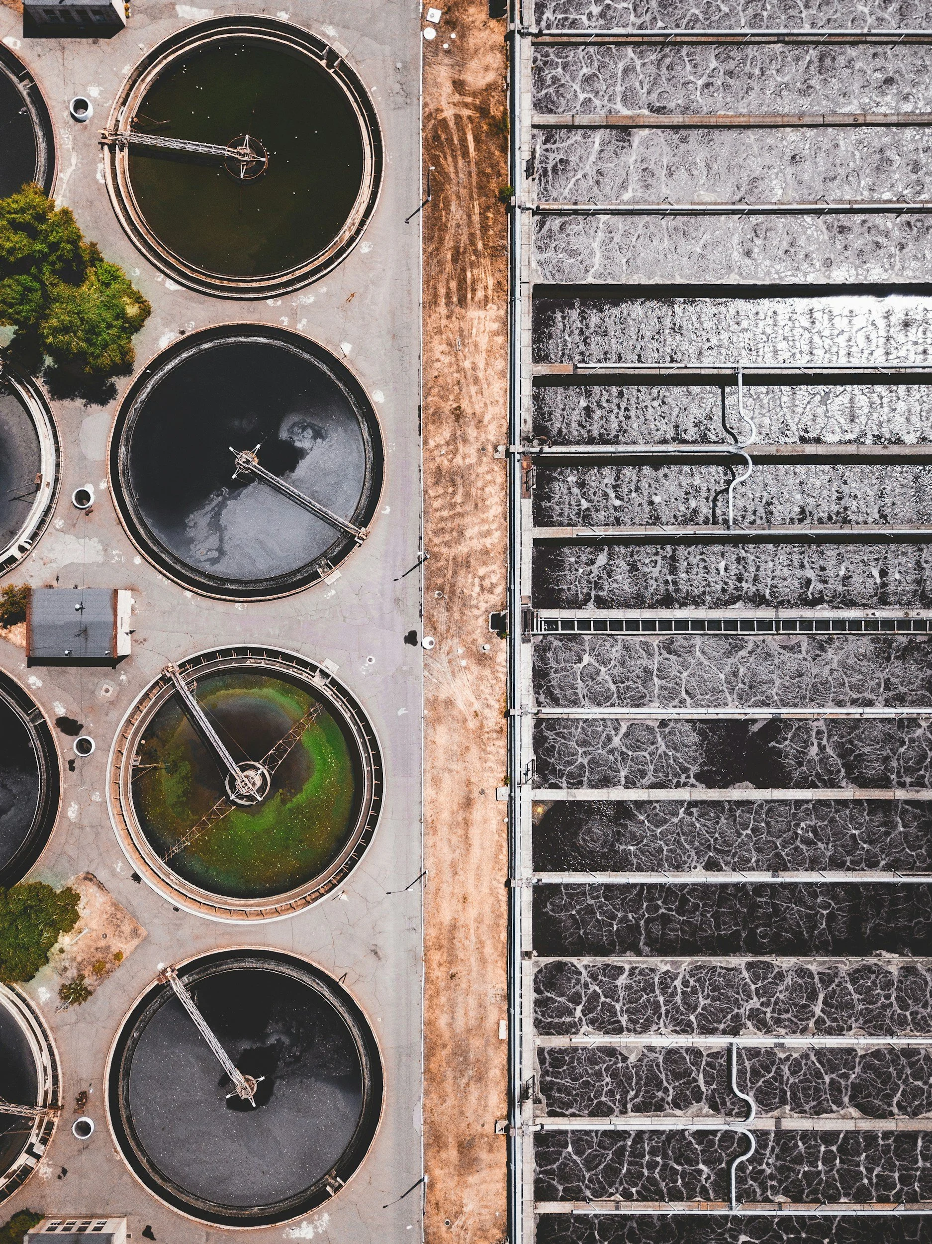 Aerial view of a water treatment plant showing four large circular tanks, separated by a concrete walkway from rectangular concrete basins with water and metal pipes.
