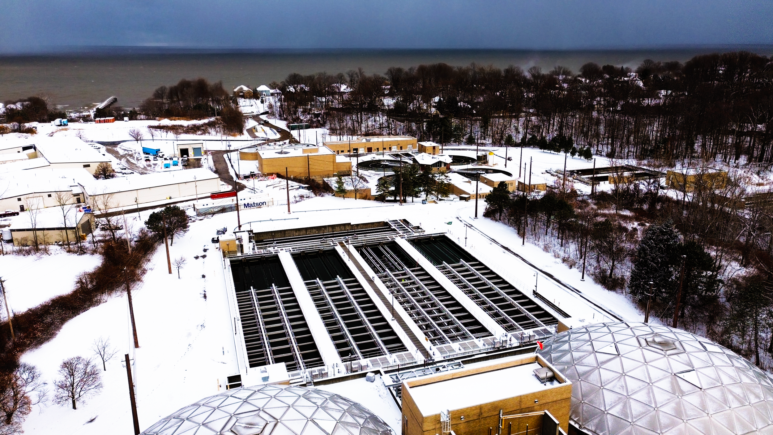 Aerial view of a snow-covered wastewater treatment plant with large circular geodesic domes and tanks, surrounded by a snowy landscape and trees, near a body of water.