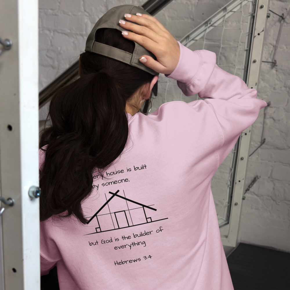 A woman wearing a pink sweatshirt with a message and a drawing of a house on the back. The message reads: "Every house is built by someone, but God is the builder of everything. Hebrews 3:4." She is touching her head with her right hand and wearing a backwards cap, standing in front of a metal structure and a white brick wall.