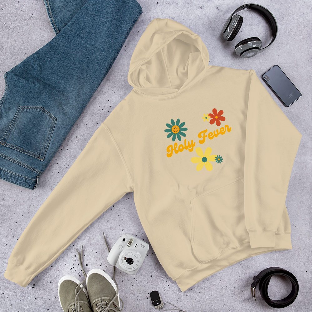 A beige hoodie with colorful daisies and the text 'Holy Fewer' written on it, laid out on a gray surface surrounded by jeans, headphones, a smartphone, a camera, a pair of shoes, a belt, and a pair of earbuds.