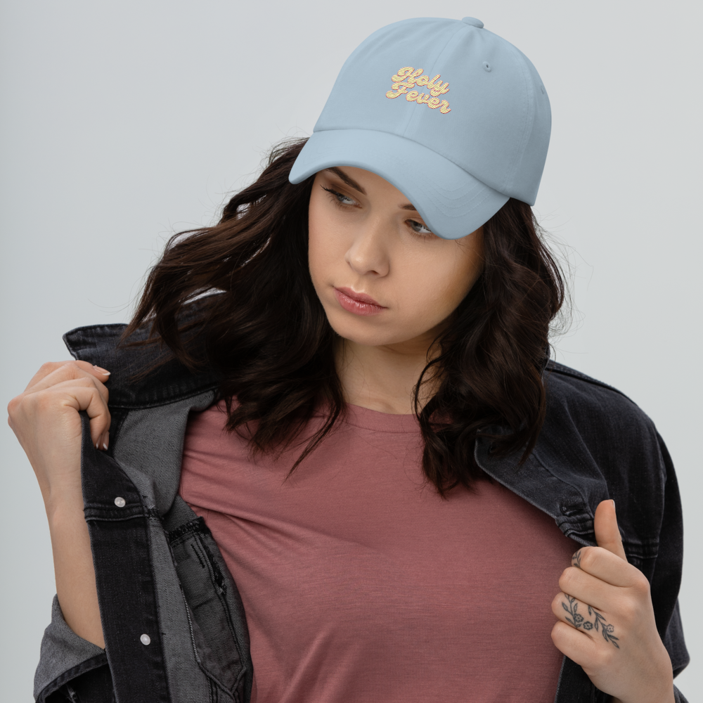 A young woman wearing a light blue baseball cap with 'Hula Flower' embroidered on it, a pink t-shirt, and a black denim jacket, looking down while holding the jacket.