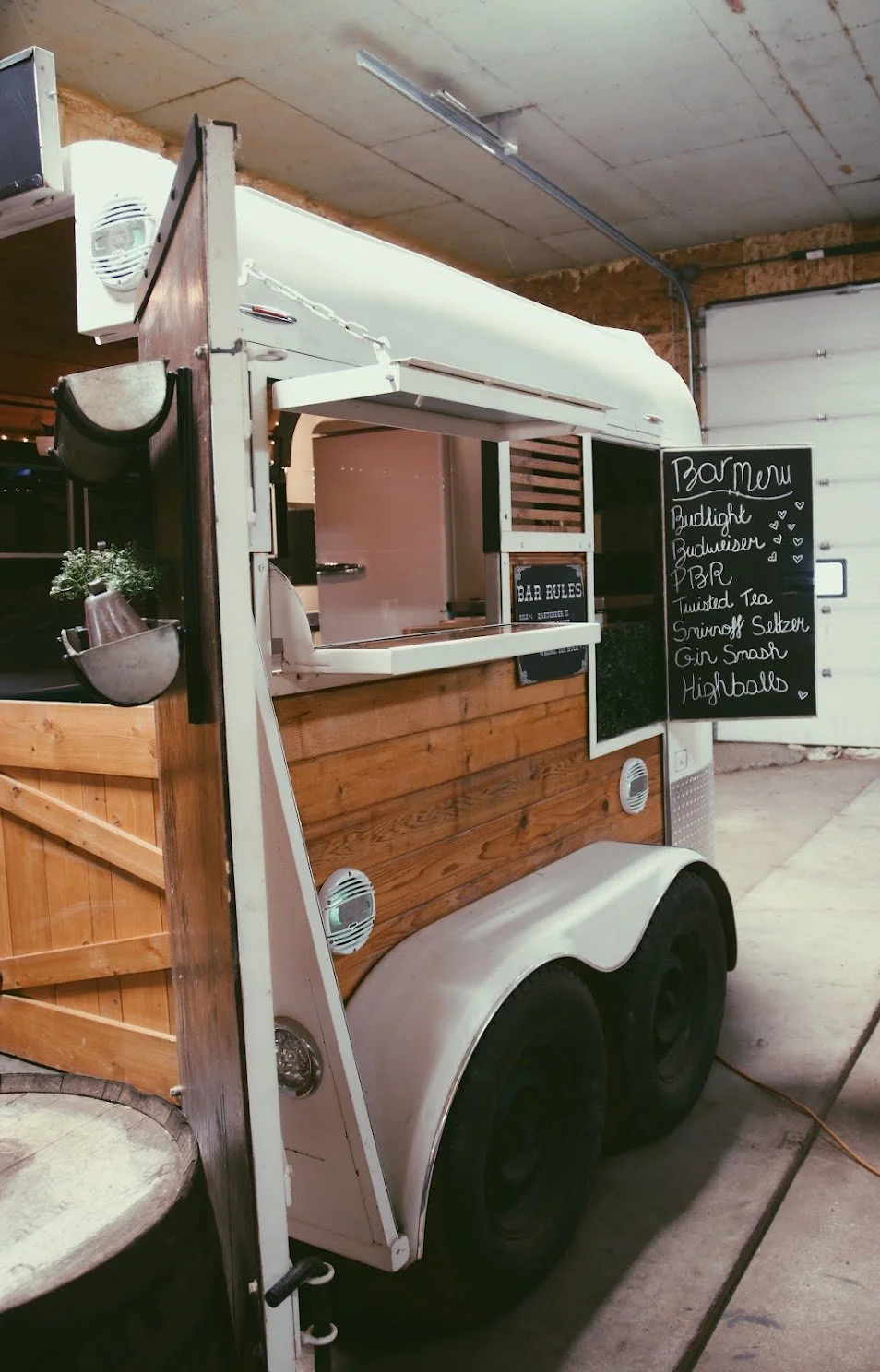 A mobile bar located inside a trailer with a chalkboard menu listing drinks like Budlight, Budweiser, cocktails, and highballs.