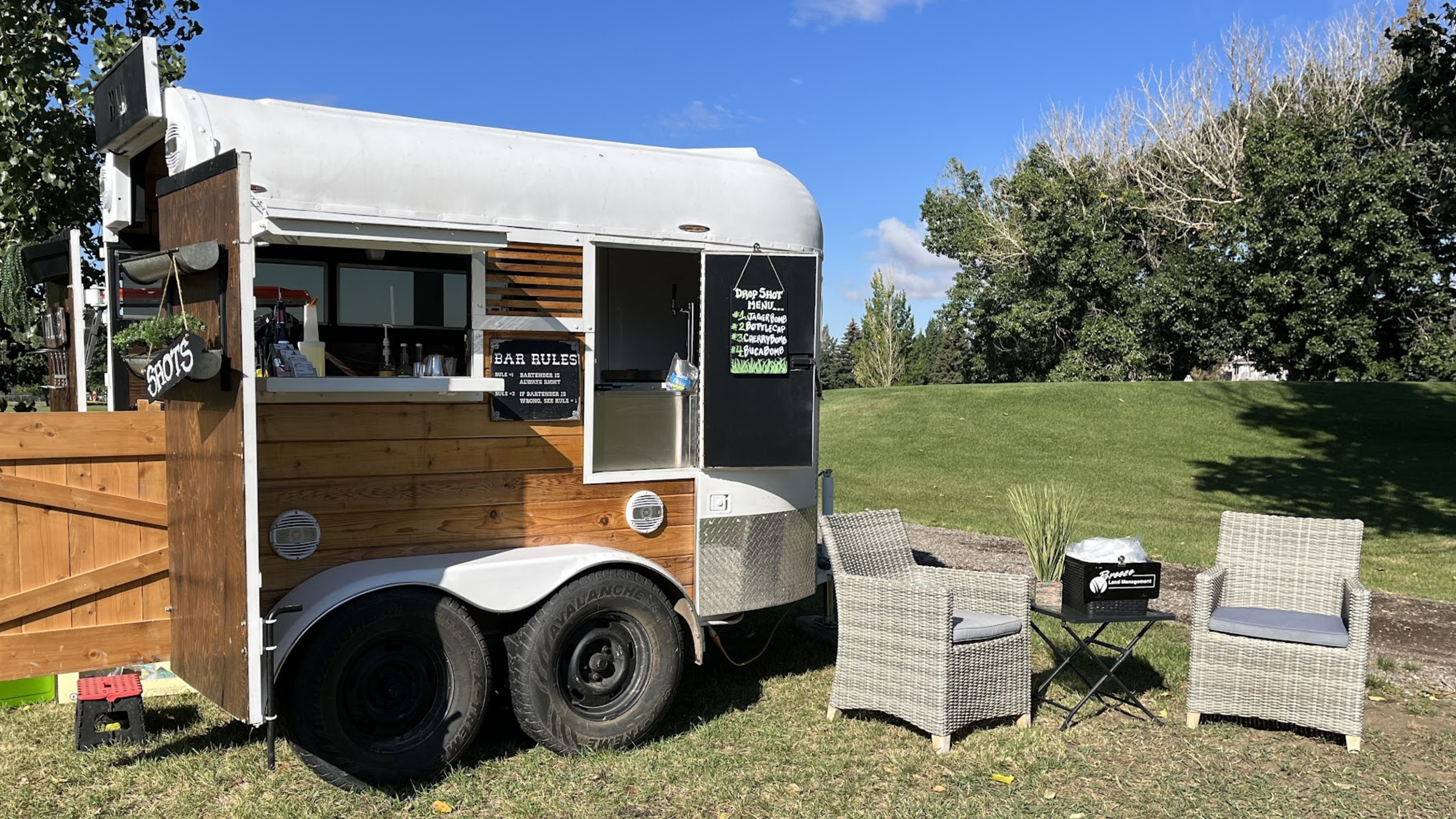 A small food trailer parked on a grassy area with two wicker chairs and a small table in front of it. The trailer has wooden paneling and a chalkboard menu on the side, with a sign that says 'SHOTS' hanging on the side. There are trees and a clear blue sky in the background.