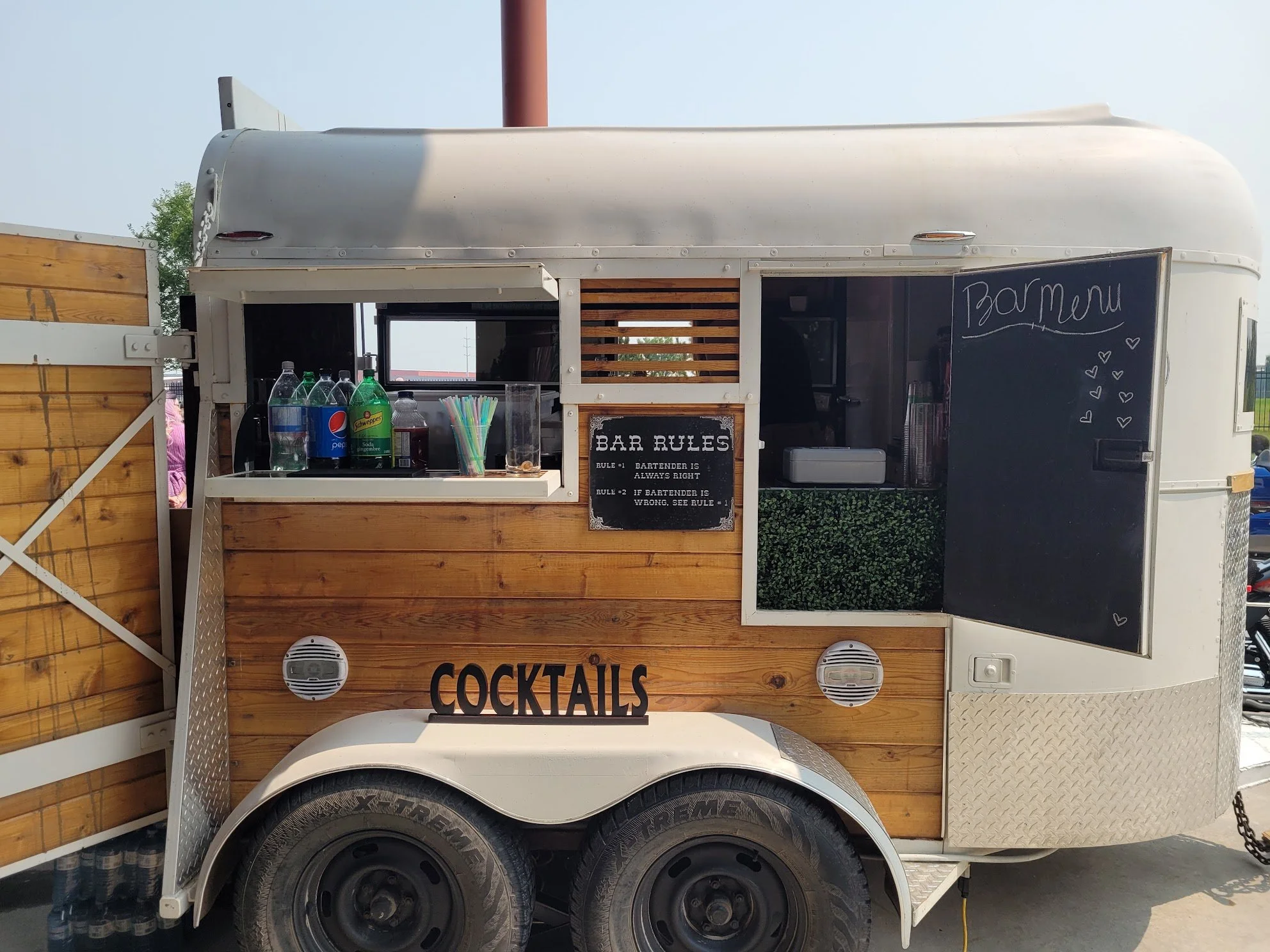 A mobile bar trailer with a wooden panel exterior, blackboard menus, and a small window with drinks and straws, set outdoors.