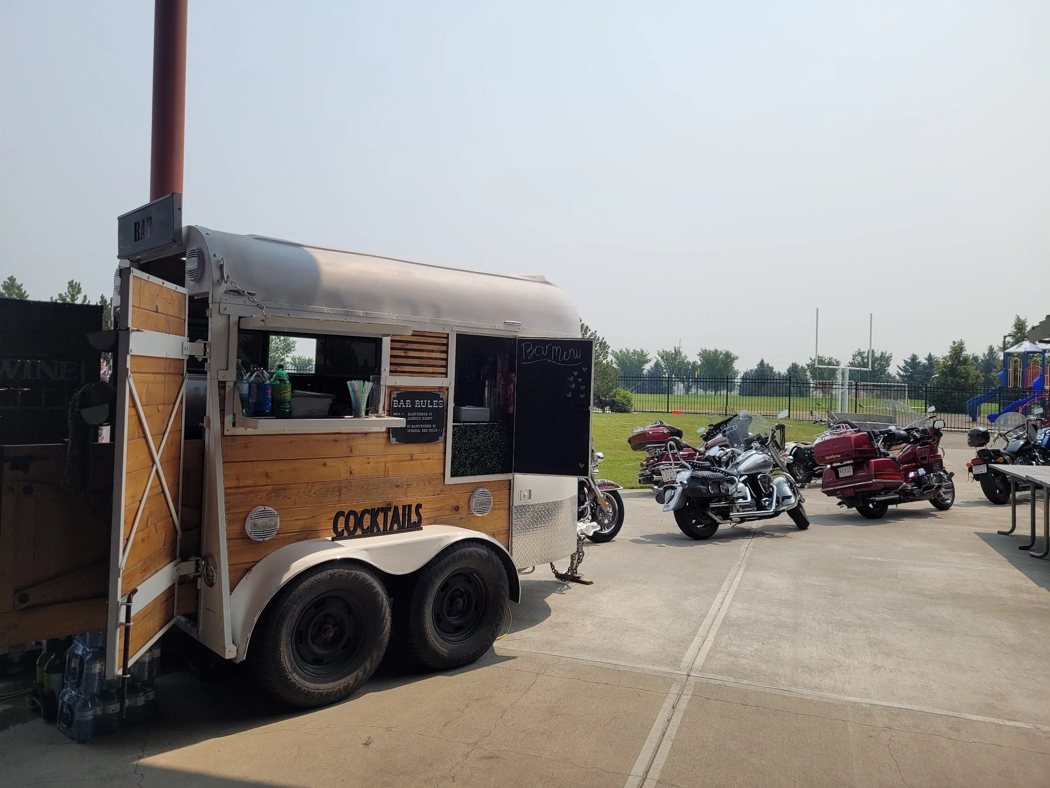Food truck selling cocktails parked outdoors near motorcycles and a playground on a sunny day.