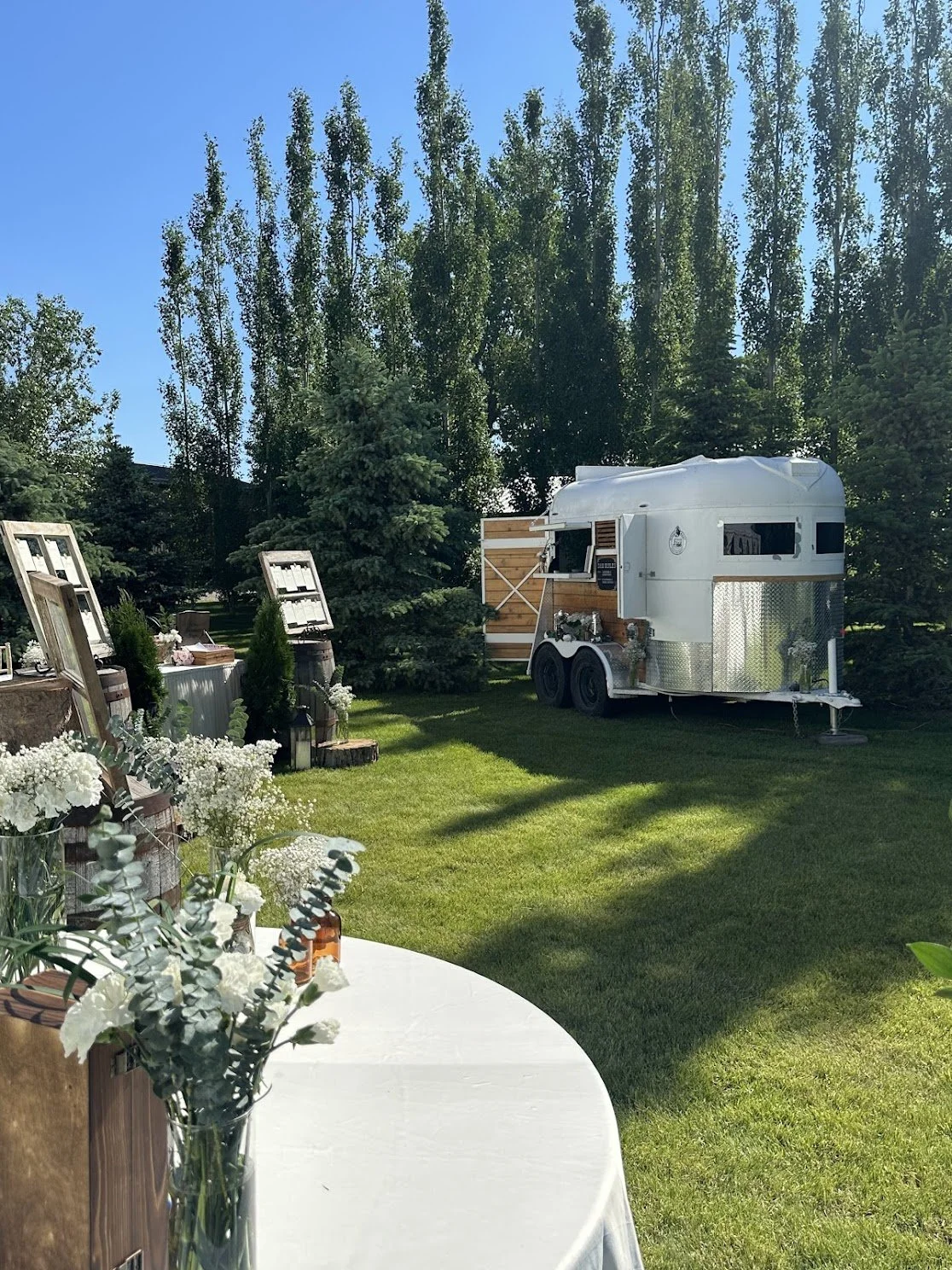 A white food truck parked on a grassy area with trees in the background and a table with flower arrangements in the foreground.