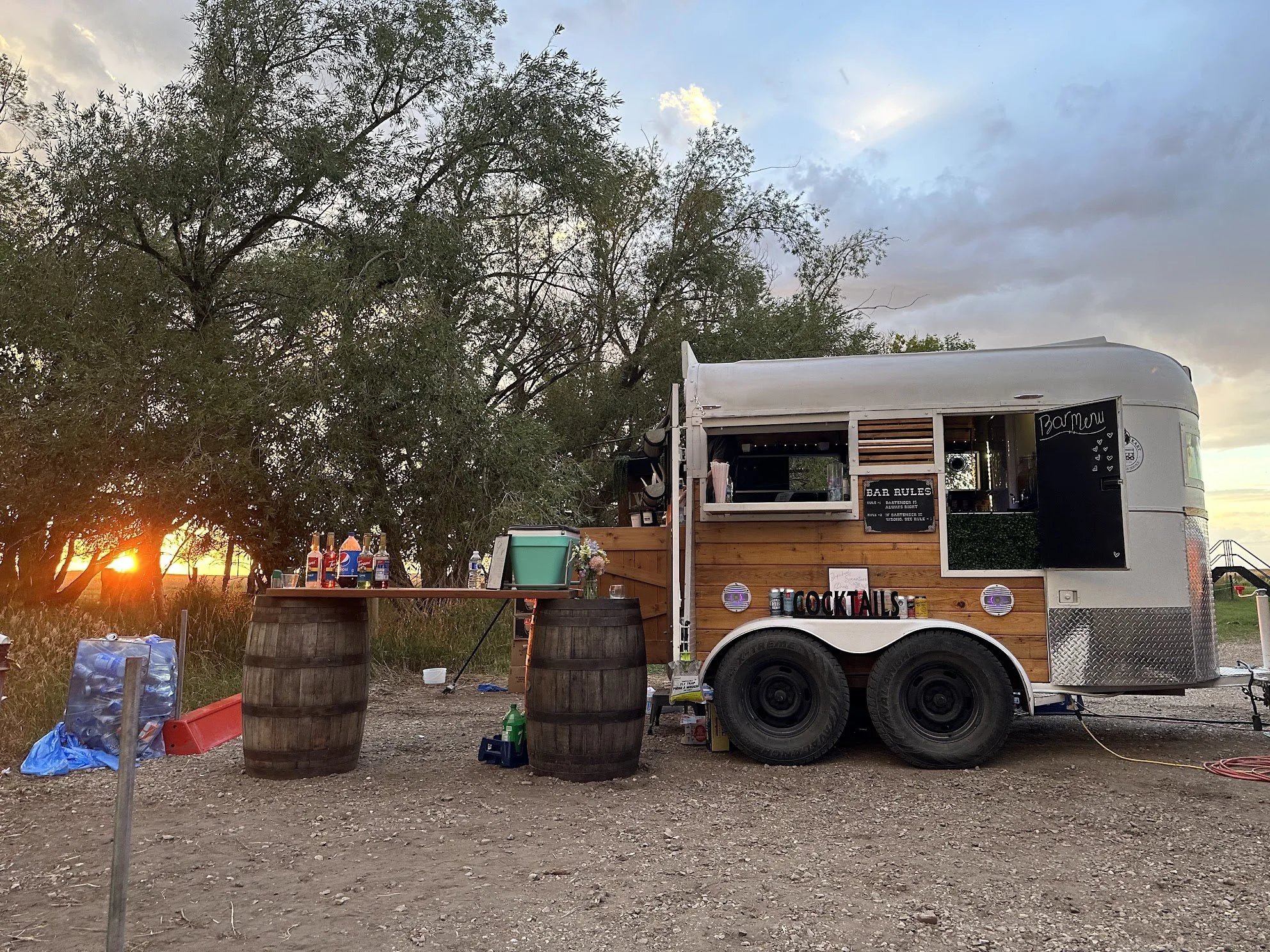 A food truck with a wood finish serving cocktails, set up outdoors at sunset near trees. It has signs for bar rules and a bar menu, with drinks and bottles displayed outside, and large barrels supporting a table with drinks and supplies.