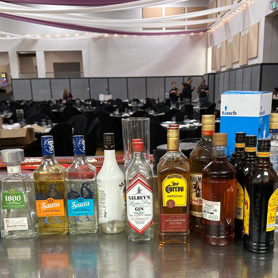 Bar with various liquor bottles, including gin, tequila, rum, and beer, set on a countertop. In the background, there is a banquet hall with round tables, black chairs, and some people standing and talking.