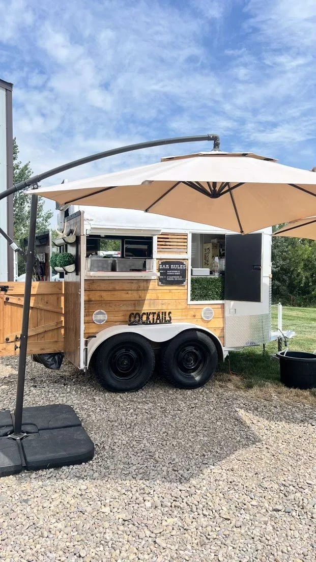 A mobile cocktail bar attached to a trailer with a large beige patio umbrella set up in front, outdoors on gravel with a clear blue sky overhead.