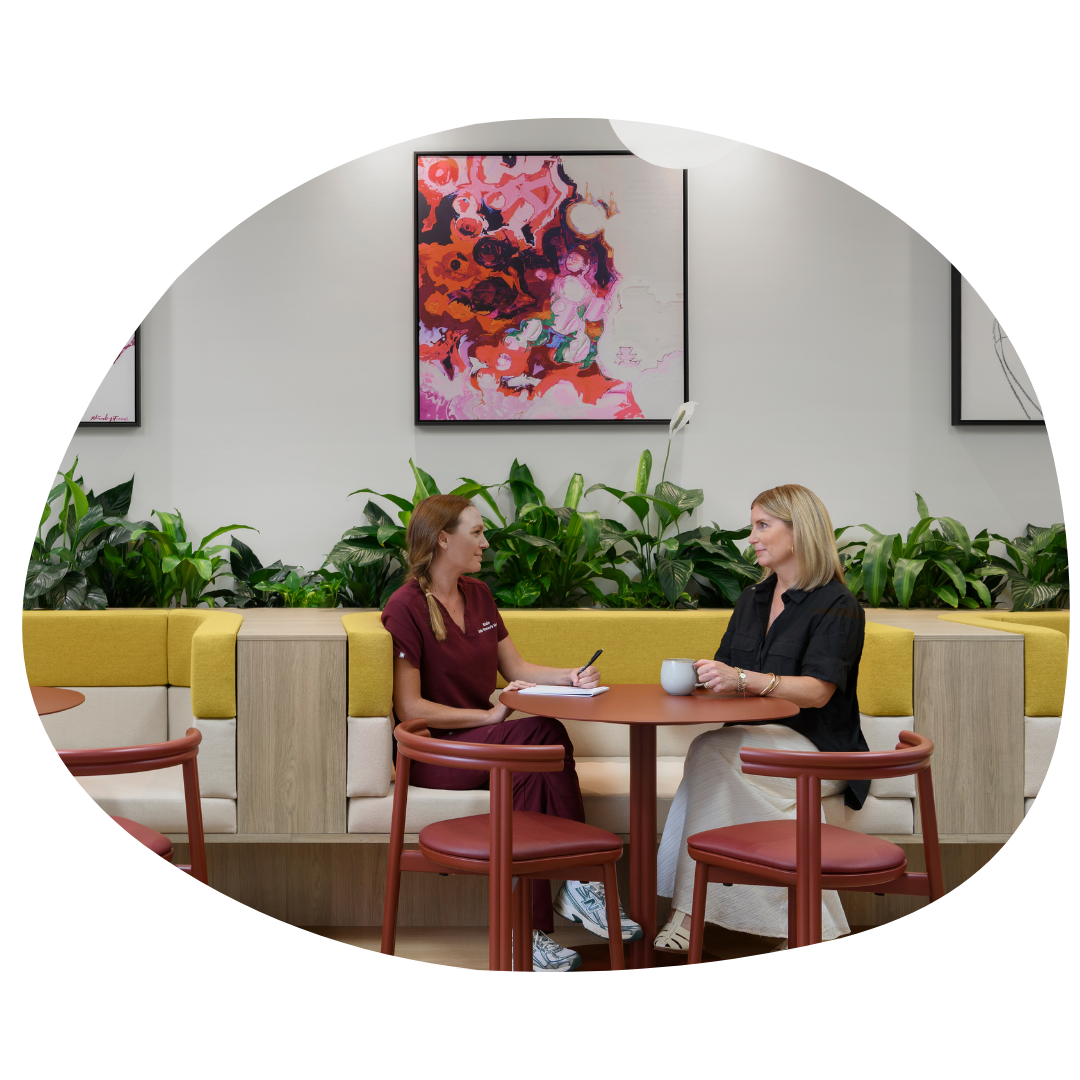 Two women sitting at a round table in a modern indoor space, engaged in a conversation. One woman is wearing a dark shirt and the other is in a maroon uniform. There are potted plants and colorful abstract paintings on the wall behind them.