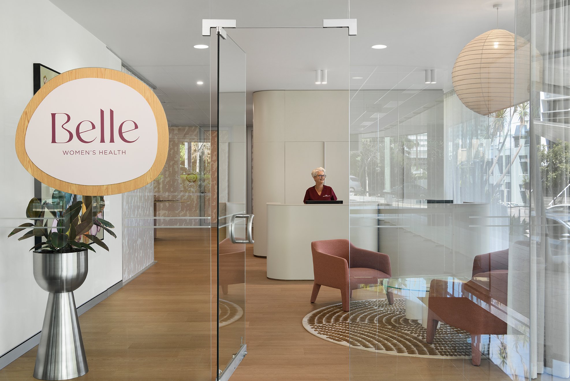 Interior of a women's health clinic with a woman receptionist standing at a curved white reception desk, glass doors, a potted plant, framed artwork on a white wall, a hanging paper lantern, and seating area with pink chairs.