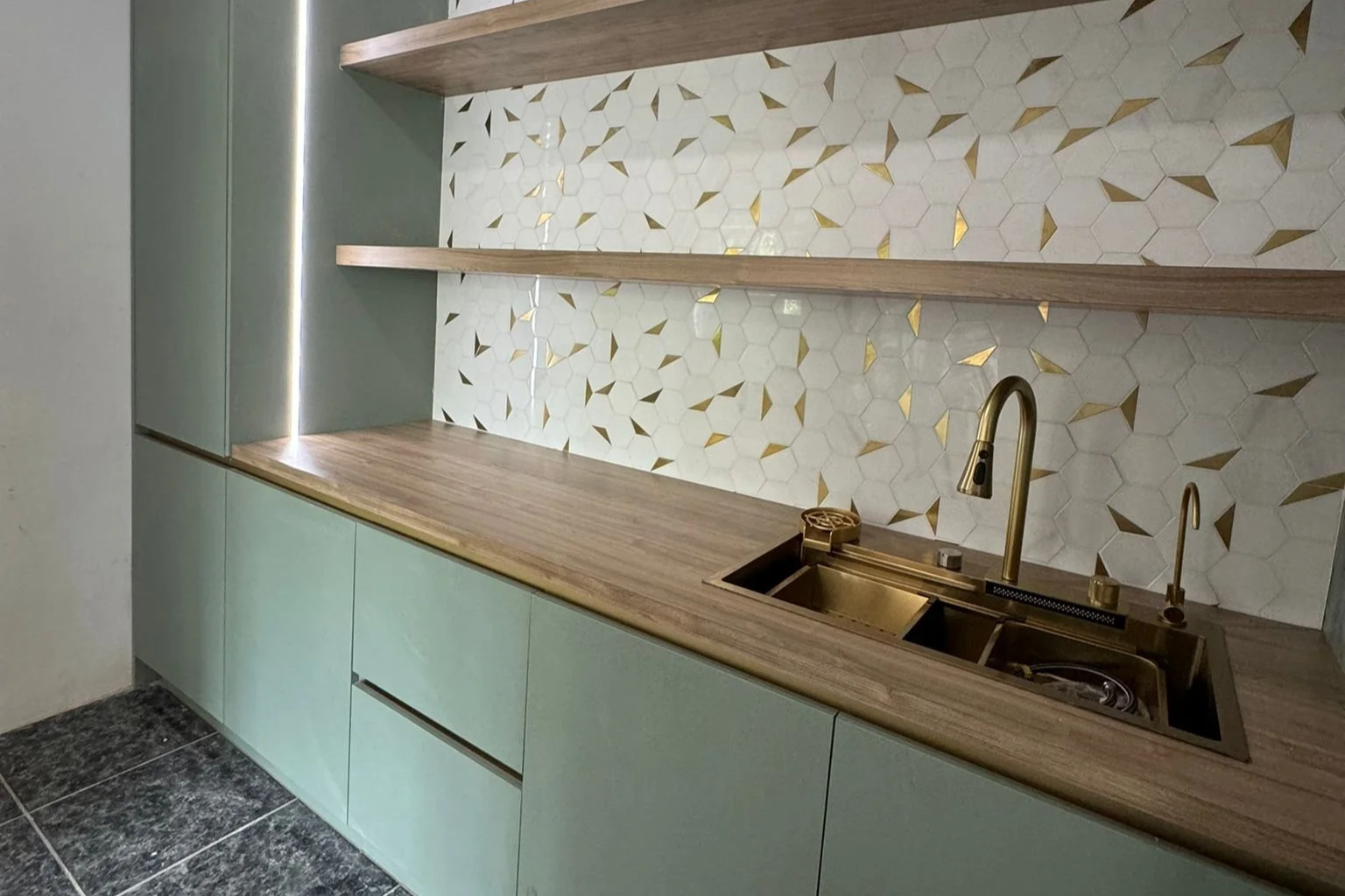 A harmonious blend of sage green cabinetry, warm wood countertops, and floating shelves against a dramatic white-and-gold hexagonal tile backsplash. Accented with elegant brass hardware and soft LED lighting, this kitchen offers a perfect balance of 