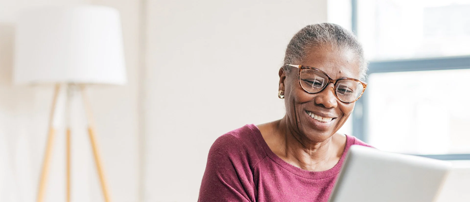 An elderly woman with gray hair, wearing glasses and a maroon shirt, smiling while looking at a laptop.