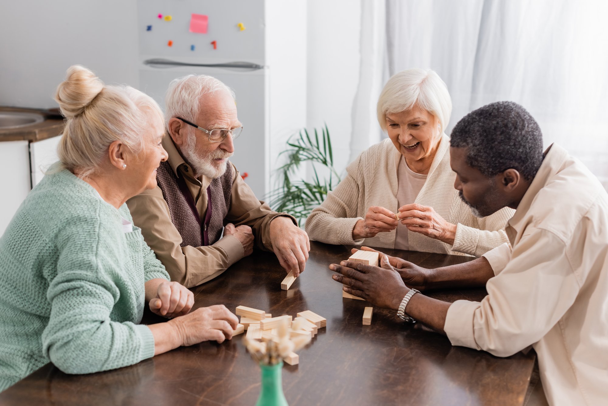 Four seniors and a caregiver playing a game with wooden blocks around a table in a living room.