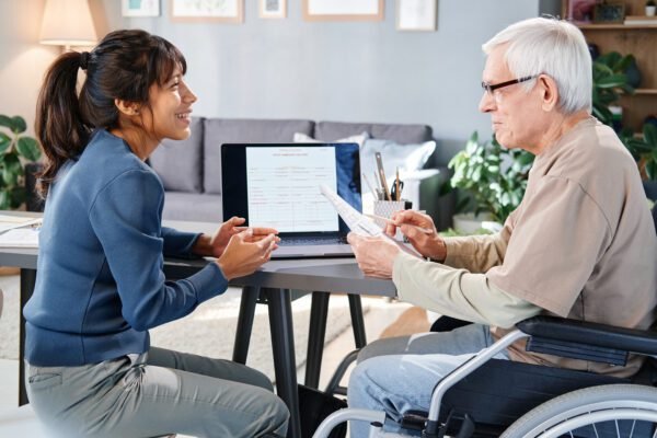 A young woman in a blue blazer talking to an elderly man in a wheelchair during a consultation at a desk with a laptop, papers, and office supplies in a bright, homey office.