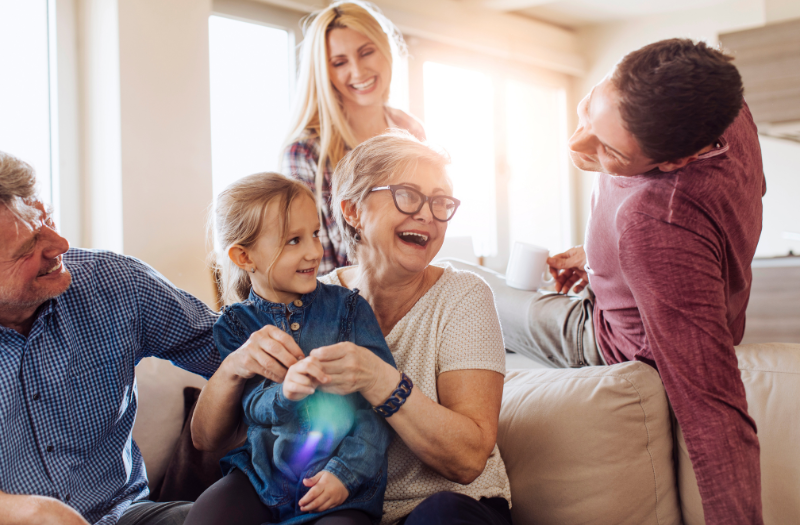 Multi-generational family sitting on a beige couch, smiling and laughing together in a bright living room.