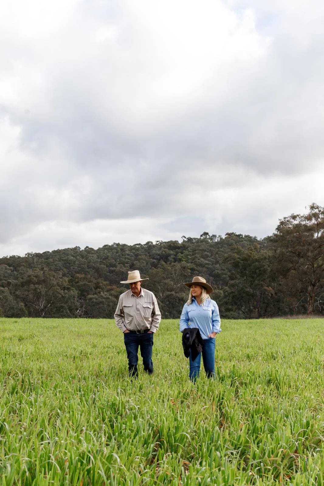 Two people walking through a green field under a cloudy sky, wearing hats and casual outdoor clothing.