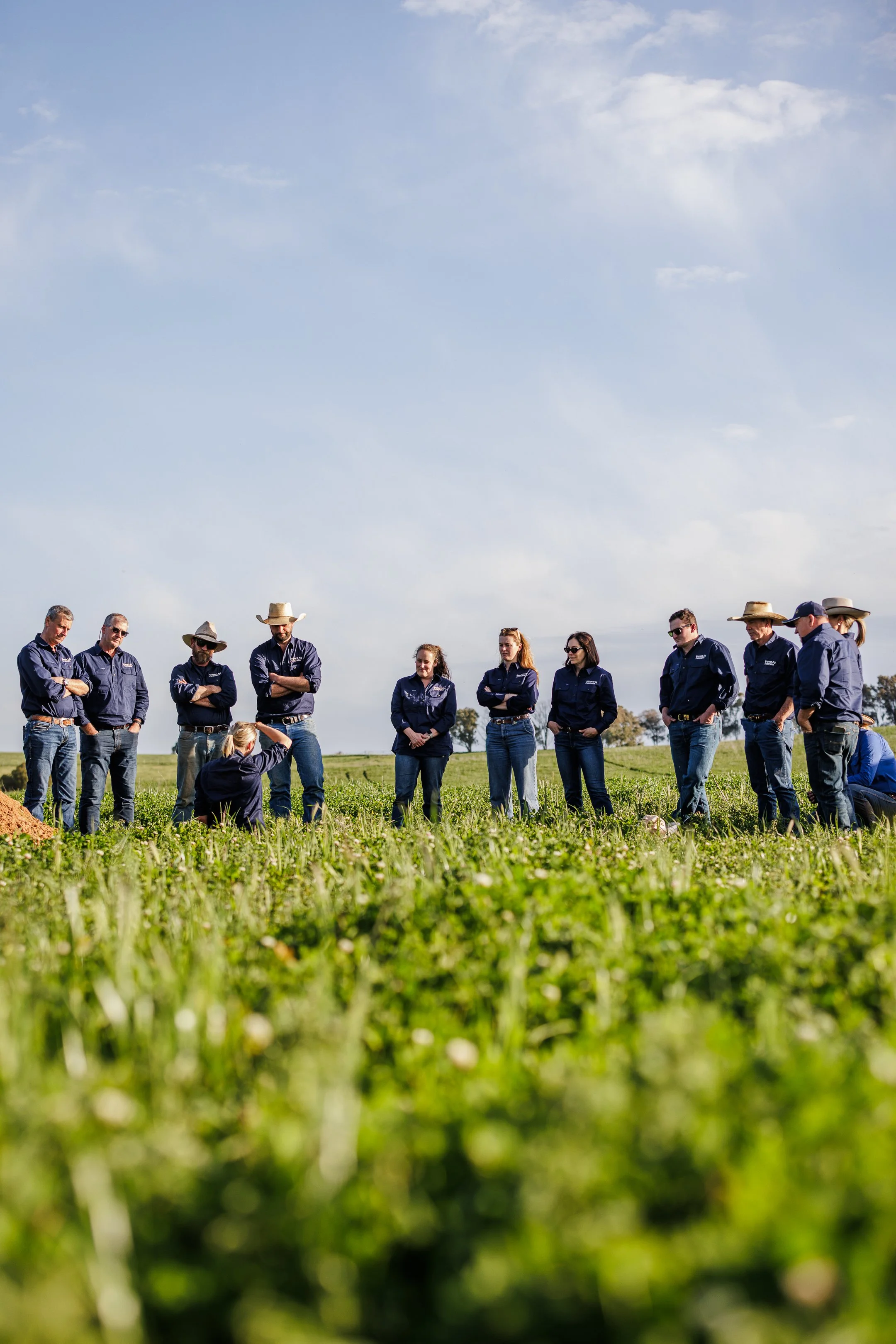 Group of people in blue shirts and jeans standing in a field, listening to a woman in a blue shirt and jeans who is demonstrating or explaining something.