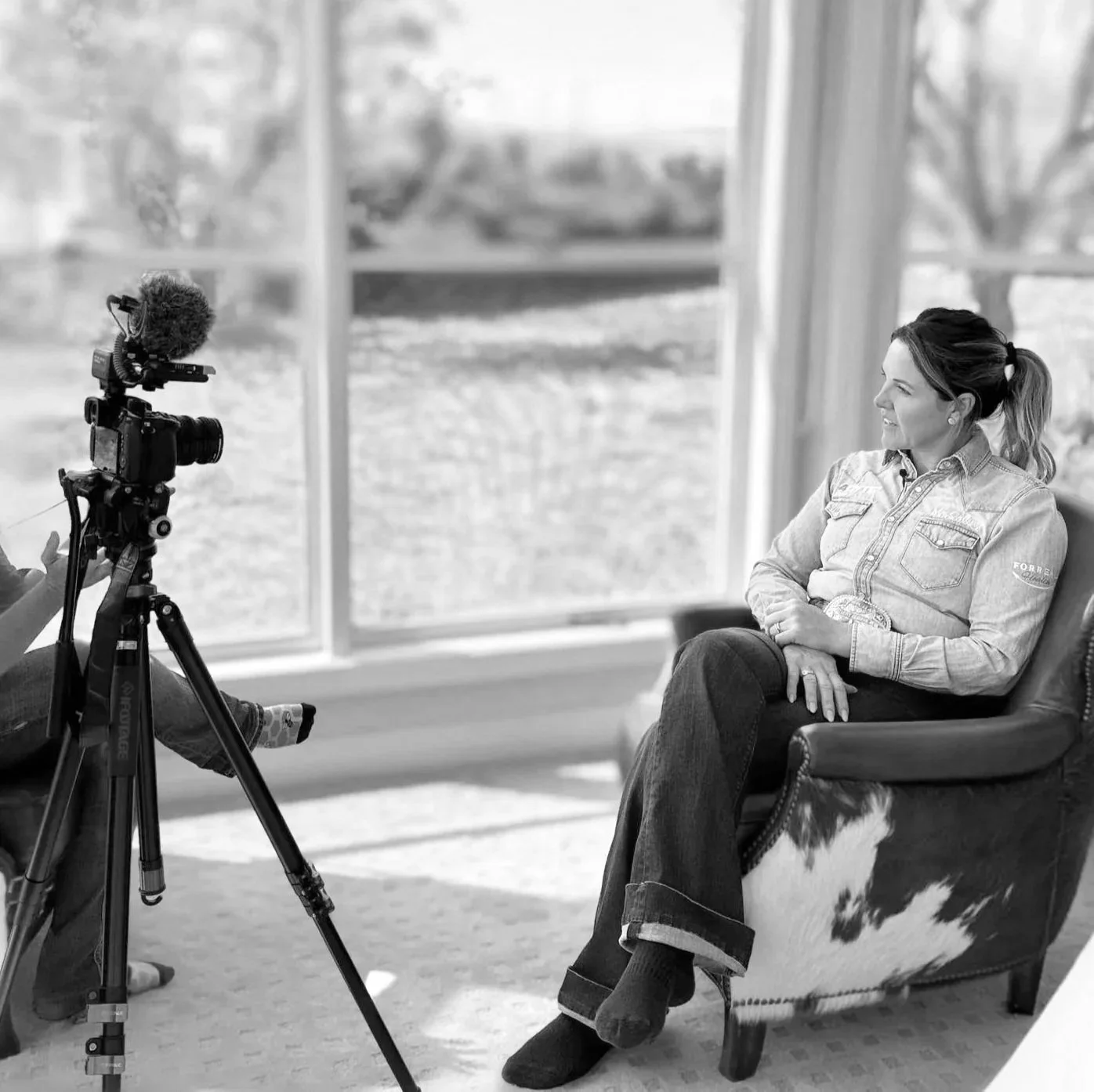 A woman with a ponytail and wearing a denim shirt and dark jeans sitting in a living room with large windows, being filmed by a person operating a camera with a microphone.