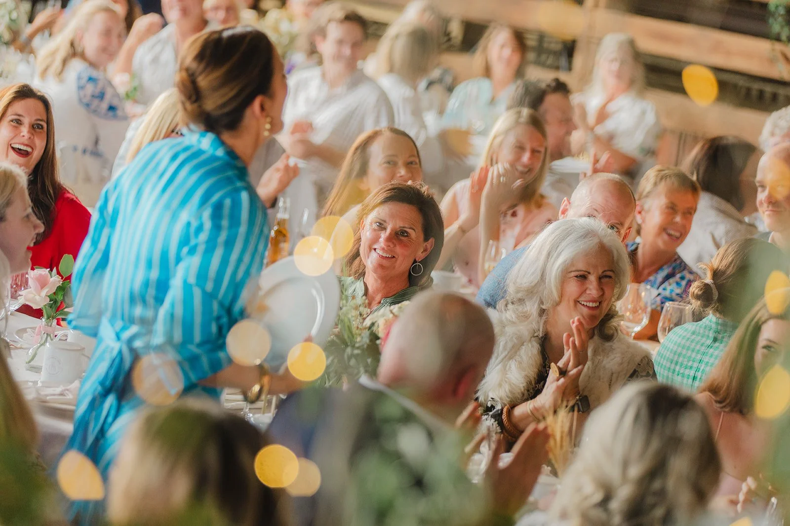 People sitting at a long table during a celebration, smiling, chatting, and enjoying the event indoors with warm lighting.