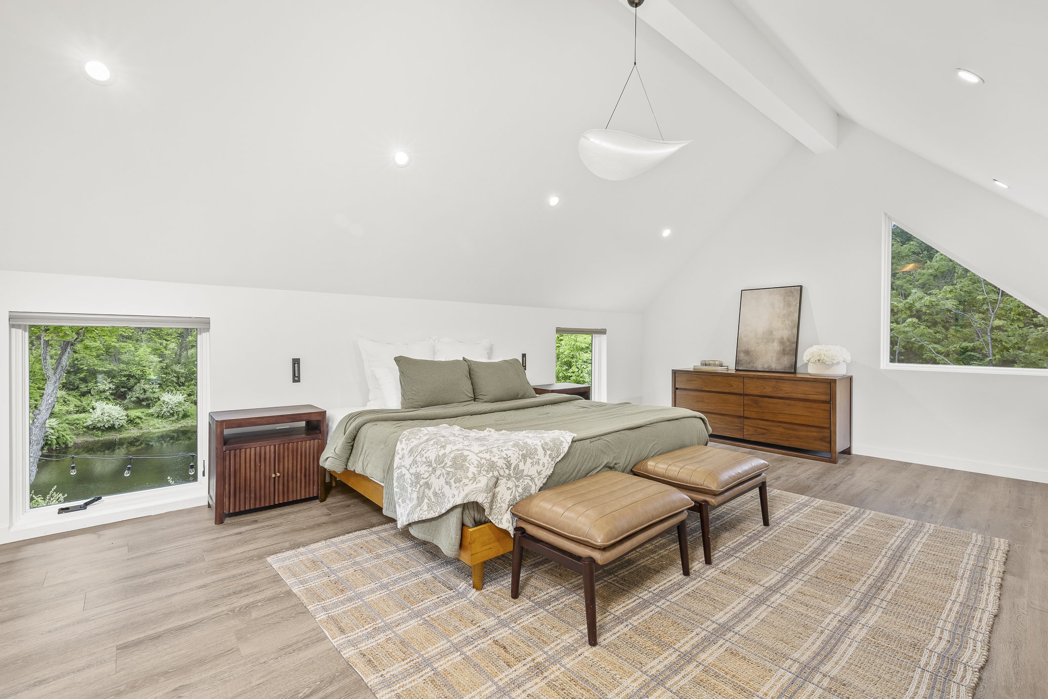 Modern bedroom with white walls and sloped ceiling, featuring a bed with green bedding, two small windows on either side, a wooden dresser with picture frame and flowers, and a large triangular window showing green trees outside.