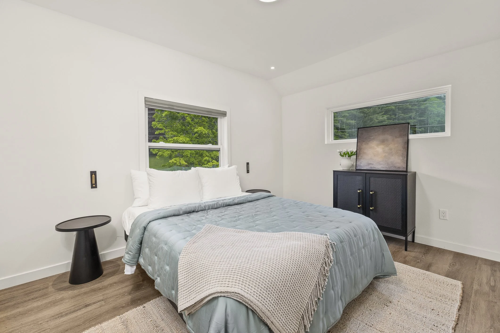 Modern minimalist bedroom with a bed, black side table, black cabinet with a plant and a TV, and large windows showing green trees outside.