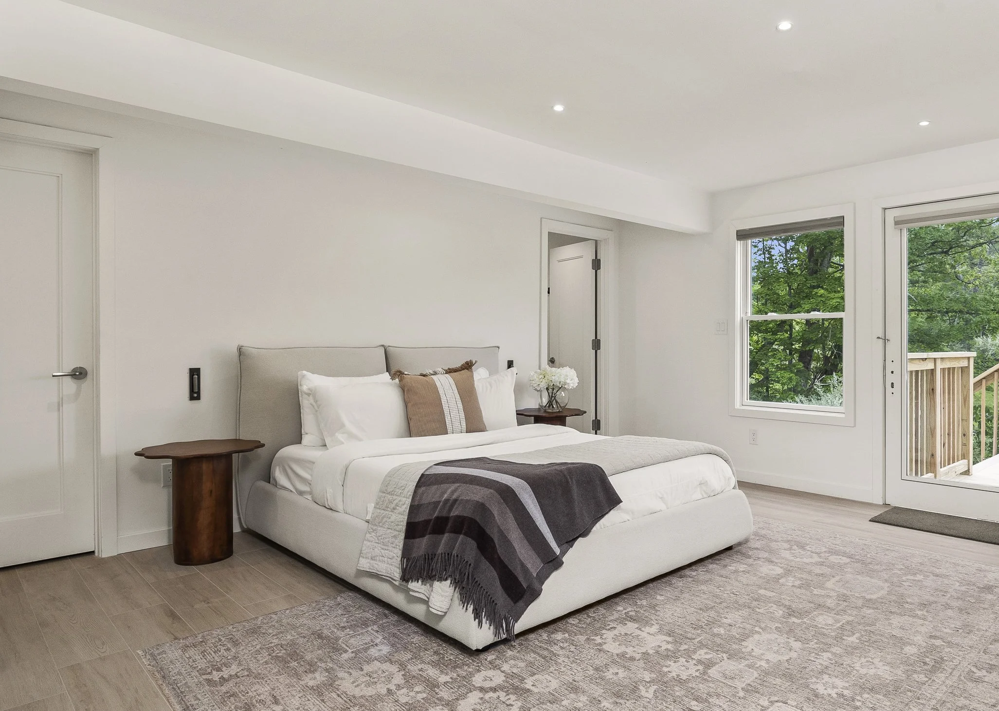 Bright, modern bedroom with a large white bed, beige headboard, and decorative pillows. There are wooden side tables with flowers and a blanket draped over the bed. Large windows show green trees outside, and there's a sliding glass door leading to a